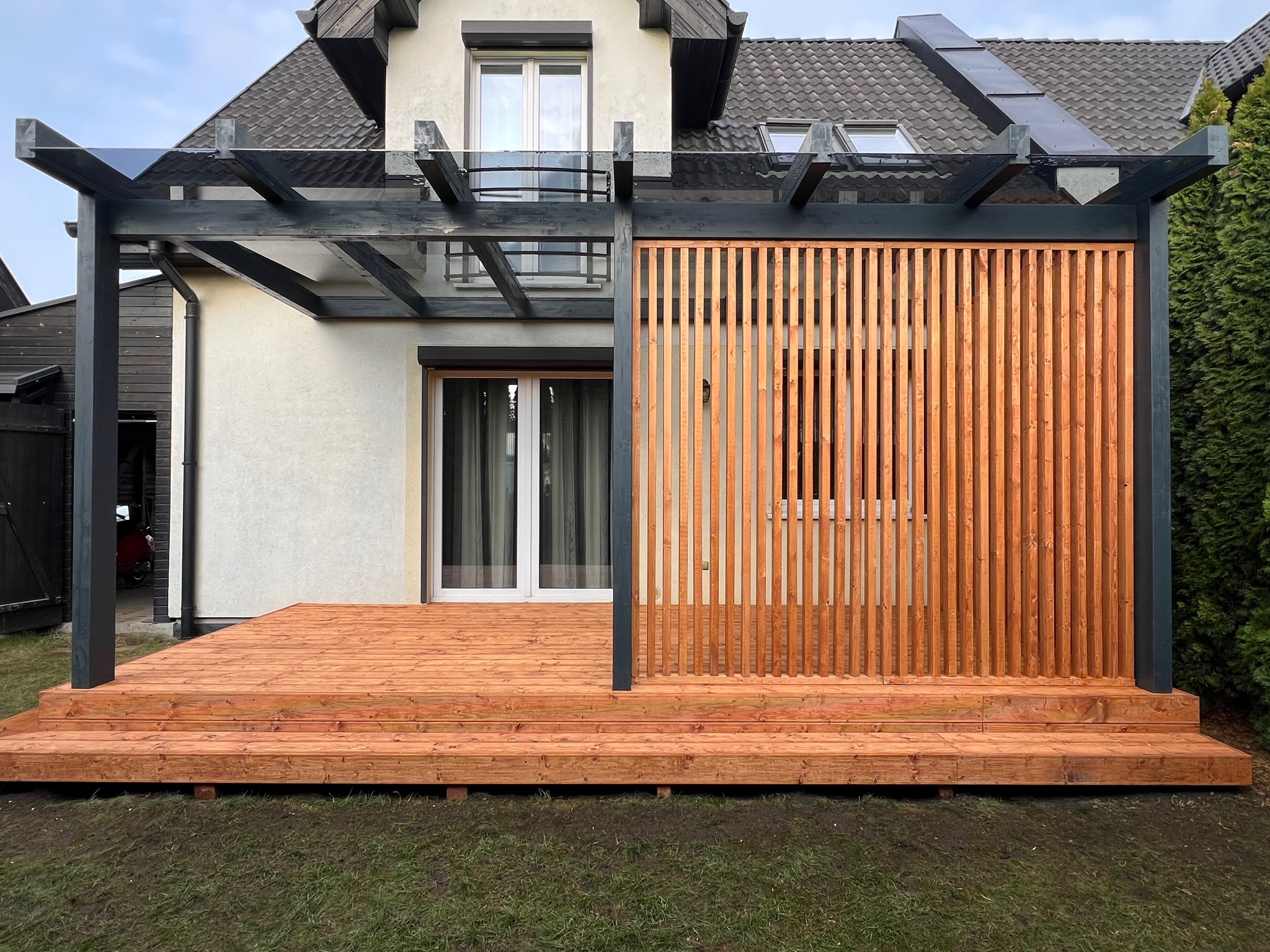 Nowoczesna pergola z drewnianą osłoną i tarasem z desek w kolorze ciepłego brązu, przylegająca do domu z jasną elewacją i ciemnym dachem.