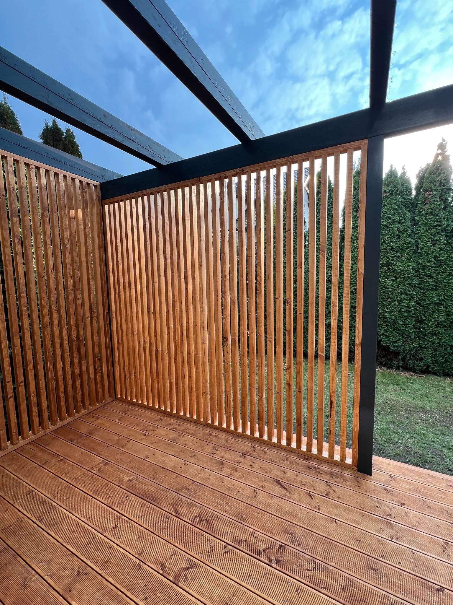 Nowoczesna, drewniana pergola z ażurowymi ściankami i tarasem z desek, na tle zielonego ogrodu i błękitnego nieba. Minimalistyczny design, naturalne materiały.