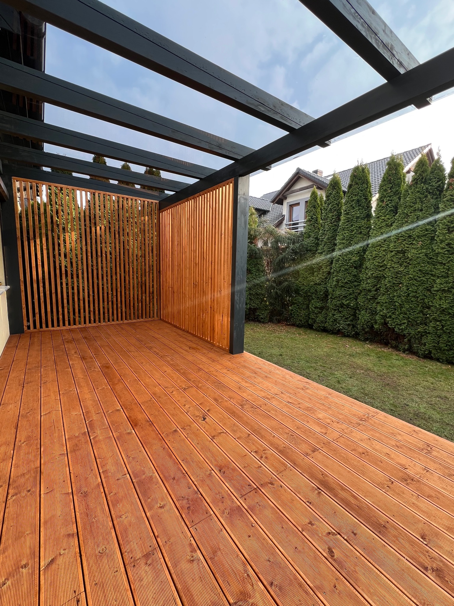 Drewniany taras z pergolą z pionowymi lamelami, widok z góry na deski, w tle zieleń i dom. Nowoczesna konstrukcja z elementami naturalnego drewna.