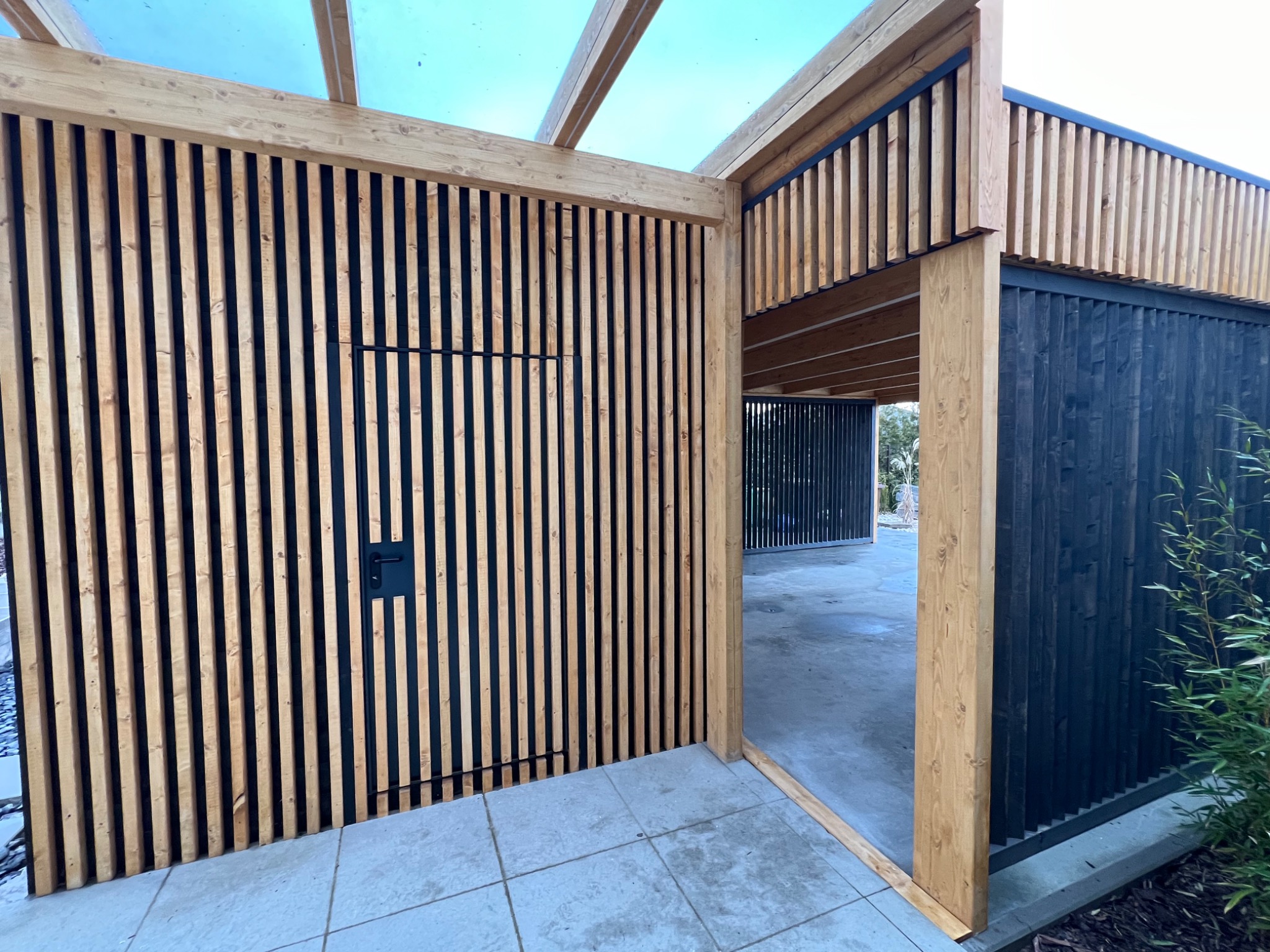 Nowoczesna konstrukcja z drewna i czarnych listew, prawdopodobnie zadaszenie tarasu lub pergola, z widocznym wejściem i fragmentem posadzki.