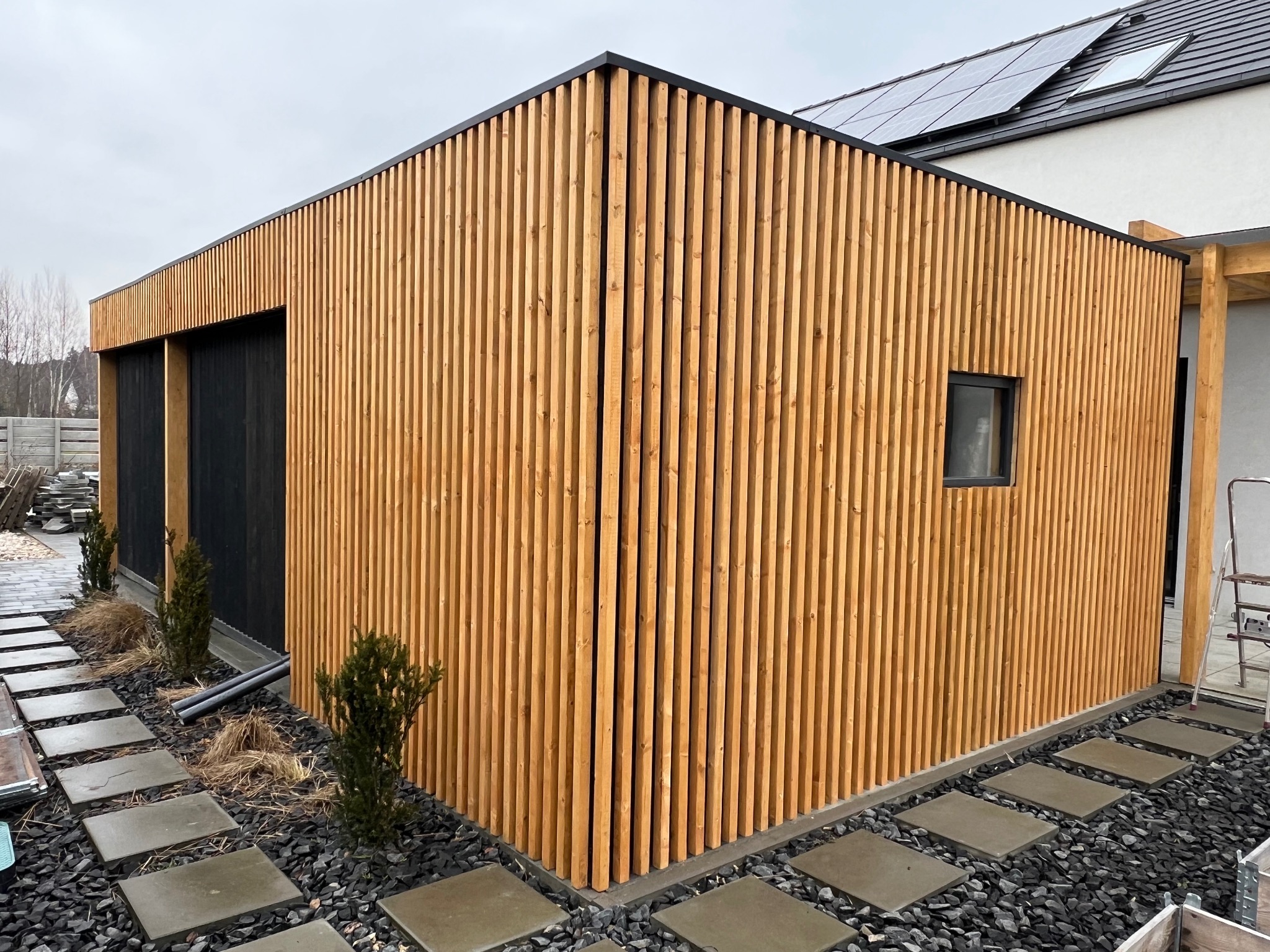 Nowoczesna, drewniana zabudowa z pionowych listew, czarne detale, kamienny chodnik i żwir. Widoczne panele słoneczne na dachu budynku w tle.