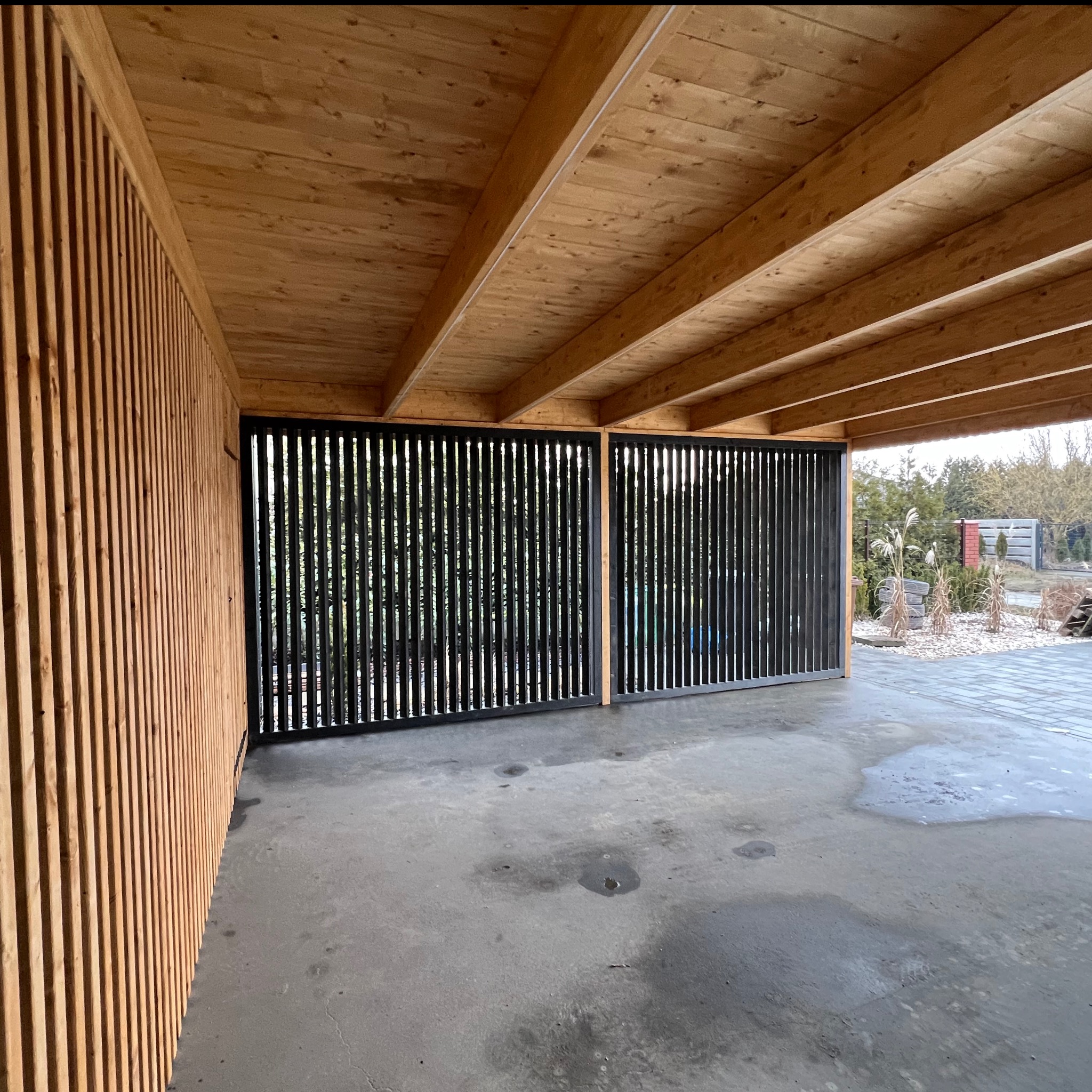 Drewniana konstrukcja z czarnymi, pionowymi panelami tworzącymi ażurową ścianę. Widoczne drewniane belki stropowe i podłoga. Element zabudowy zewnętrznej.