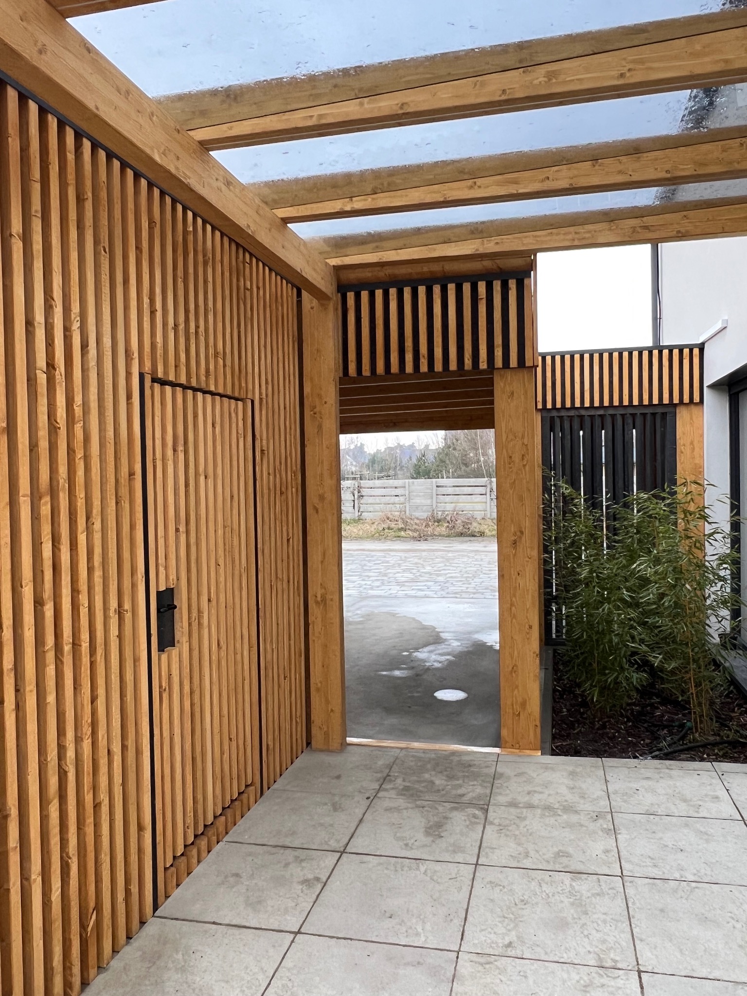 Drewniana pergola z przeszklonym dachem i ścianą z pionowych listew z ukrytymi drzwiami, widok z zewnątrz na wyłożony płytami taras.
