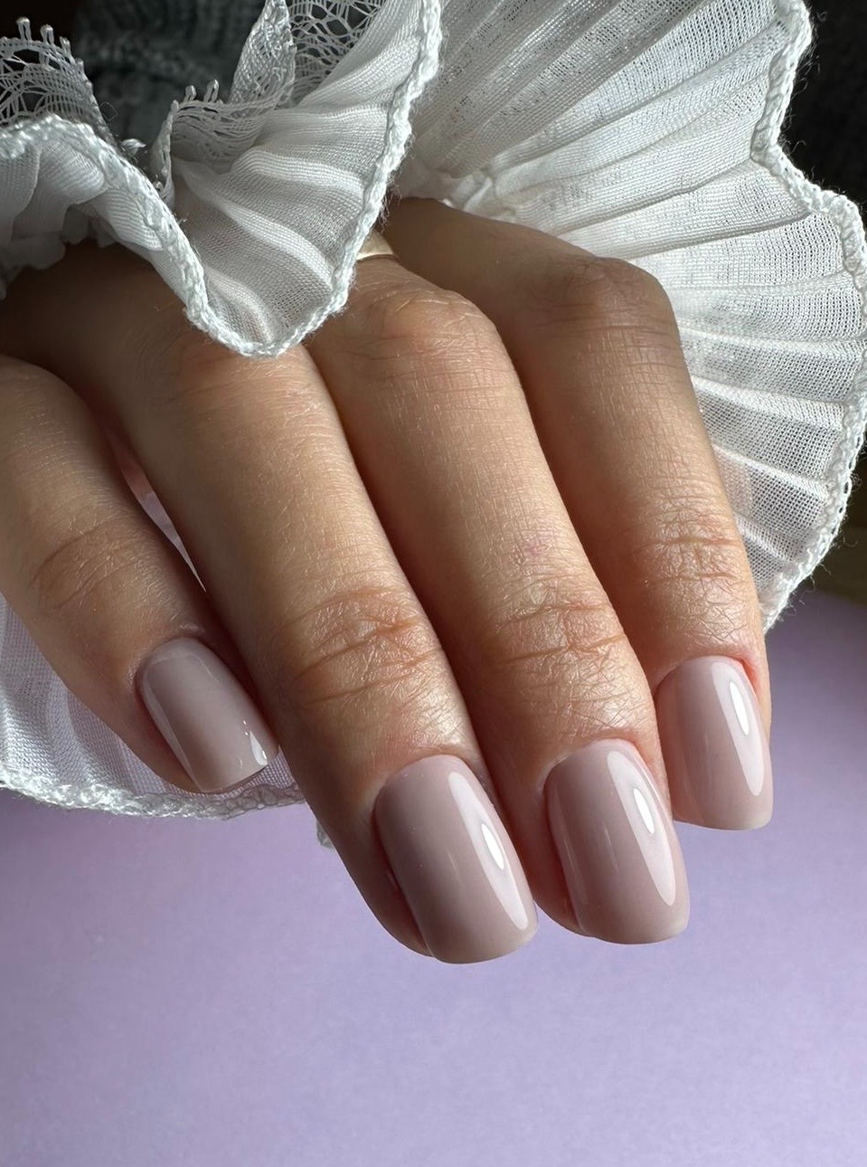 Delikatne, beżowe paznokcie kwadratowe z subtelnym połyskiem, wykonane w Bytomiu, na tle fioletowego blatu i białej, plisowanej falbanki.