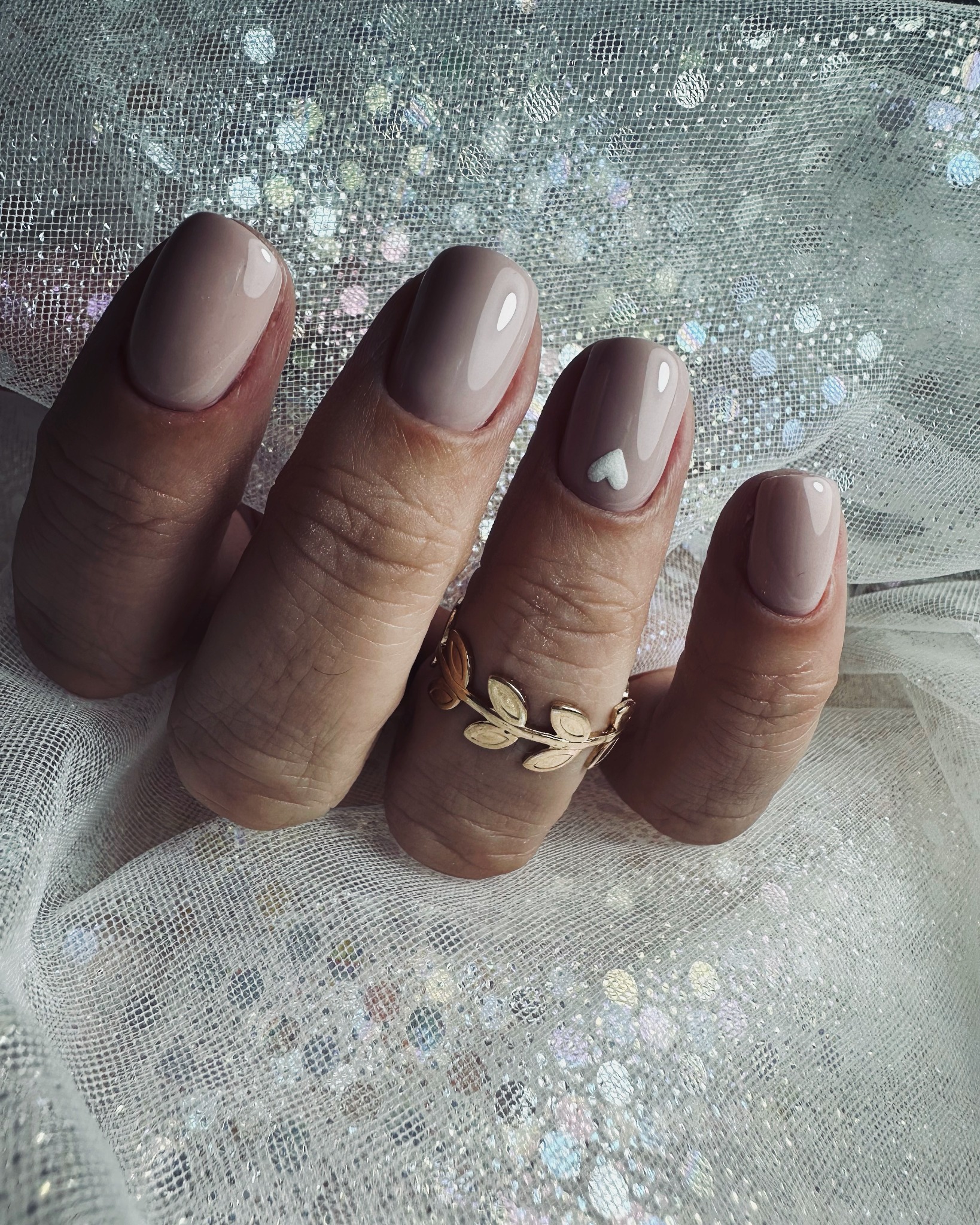 Paznokcie w odcieniu nude z małym białym serduszkiem na jednym palcu, złoty pierścionek z motywem liści na tle błyszczącej, srebrnej tkaniny.