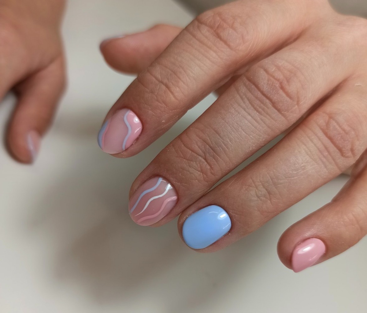 Dłonie z pomalowanymi paznokciami w pastelowych kolorach: jasny róż i błękit, zdobione delikatnymi, falistymi paskami.