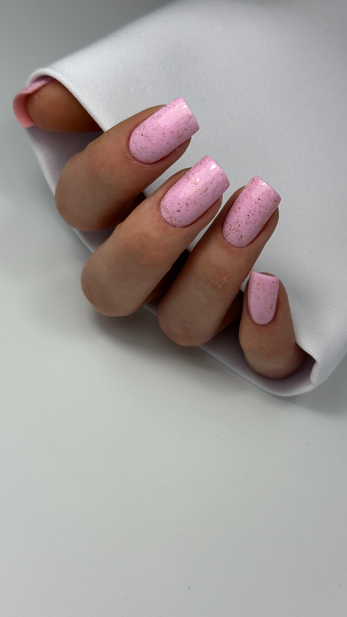 Ręka z kwadratowymi paznokciami pomalowanymi na różowo z delikatnymi, złotymi drobinkami, spoczywająca na białej serwetce.