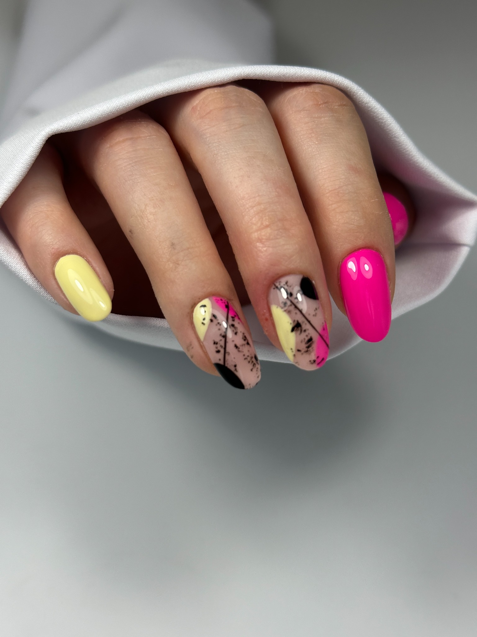 Dłoń z abstrakcyjnym zdobieniem paznokci: żółty, różowy i czarny na beżowym tle. Jasne, neonowe kolory lakieru i geometryczny wzór. Część białej koszuli w tle.