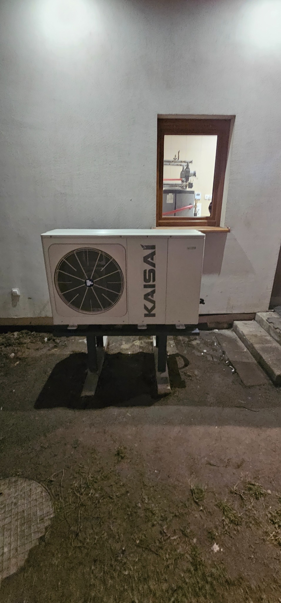Biała pompa ciepła marki Kaisai zamontowana na zewnątrz budynku, widok przez okno na instalację grzewczą wewnątrz.