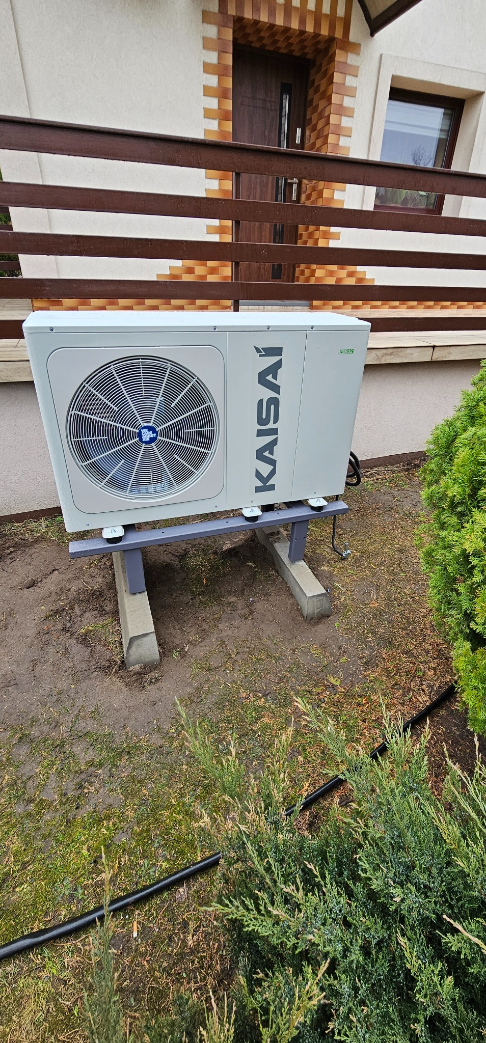 Zewnętrzna jednostka pompy ciepła Kaisai, umieszczona na szarym stelażu z betonowymi podstawami, obok zielonego krzewu i czarnego węża, na tle elewacji domu z drewnianą balustradą.