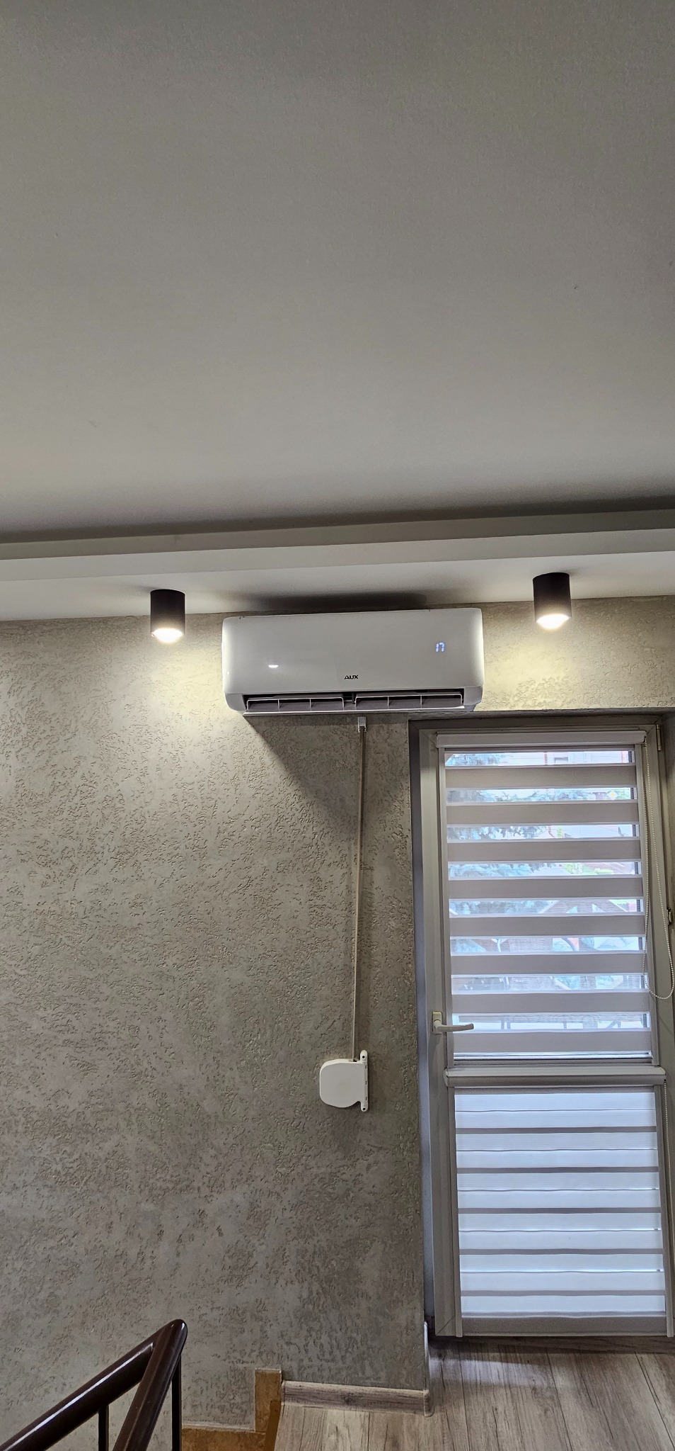 Biały klimatyzator marki AUX zamontowany na ścianie o fakturze baranka, widoczny termostat wskazujący temperaturę 17 stopni, obok okno z roletami dzień-noc.
