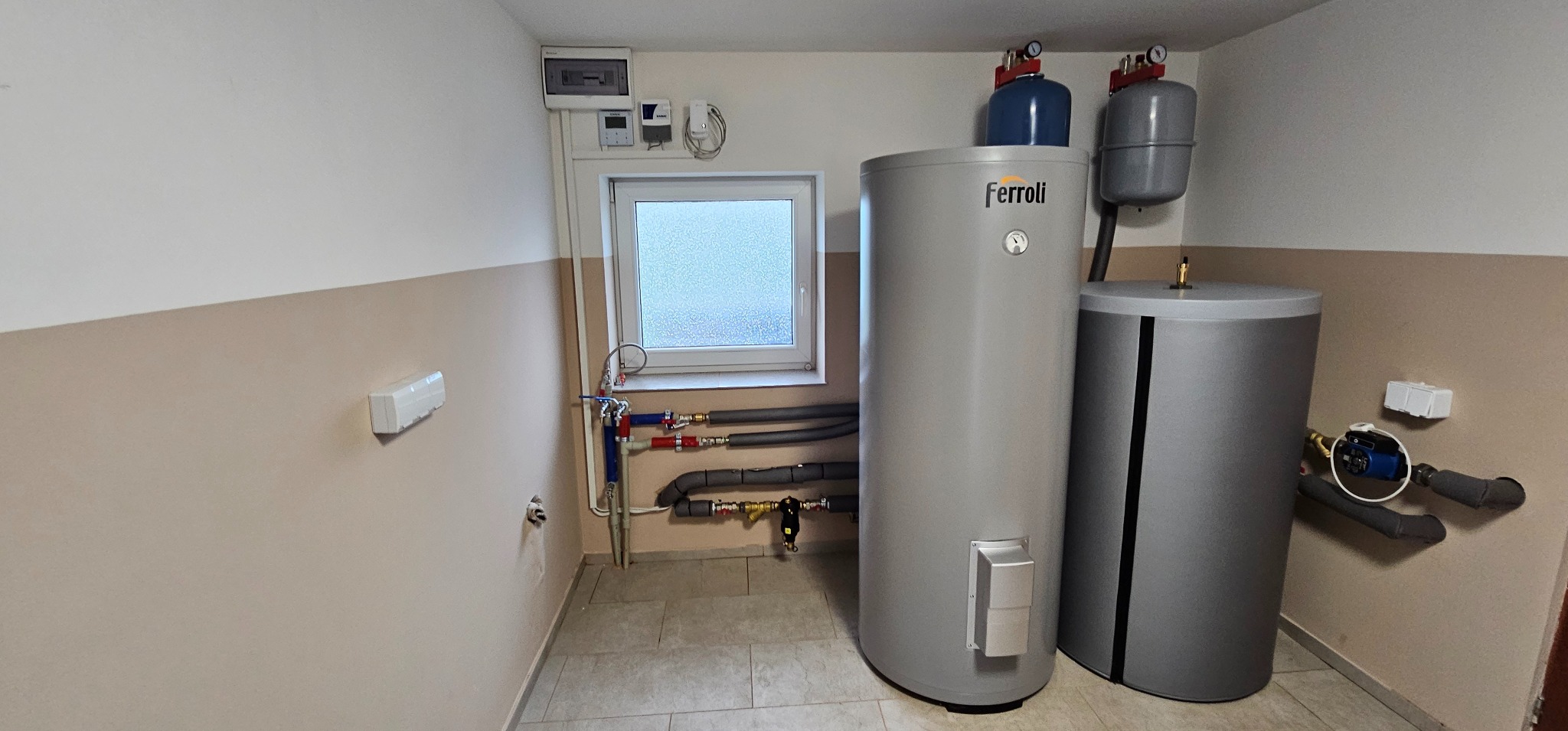 Dwa szare zbiorniki Ferroli w pomieszczeniu z oknem i instalacją hydrauliczną, widoczne także zabezpieczenia i sterowniki na ścianie.
