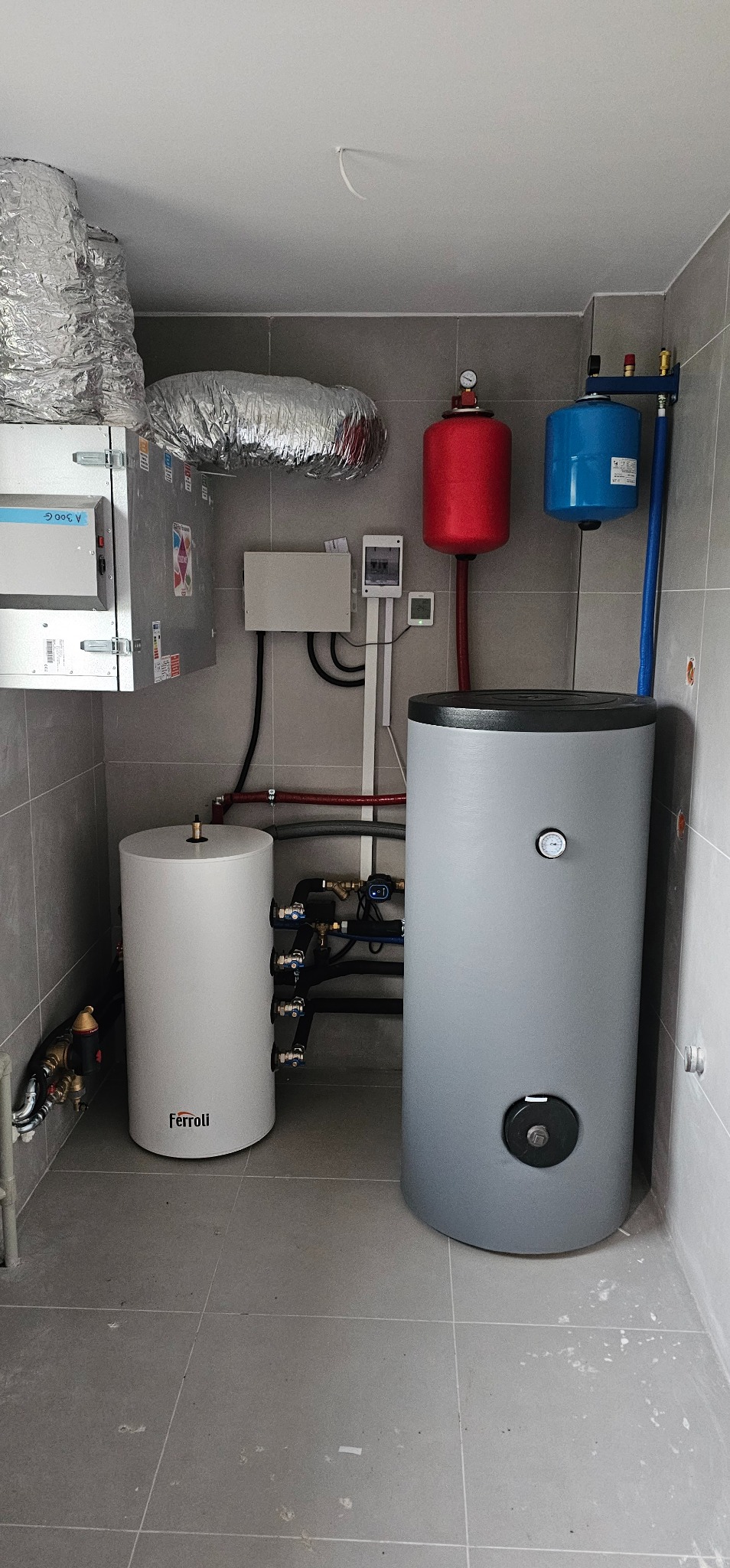 W kotłowni zainstalowano biały zasobnik ciepłej wody użytkowej Ferroli i szary zbiornik buforowy, połączone rurami. Nad nimi widoczne są czerwony i niebieski zbiornik przeponowy oraz kanały...