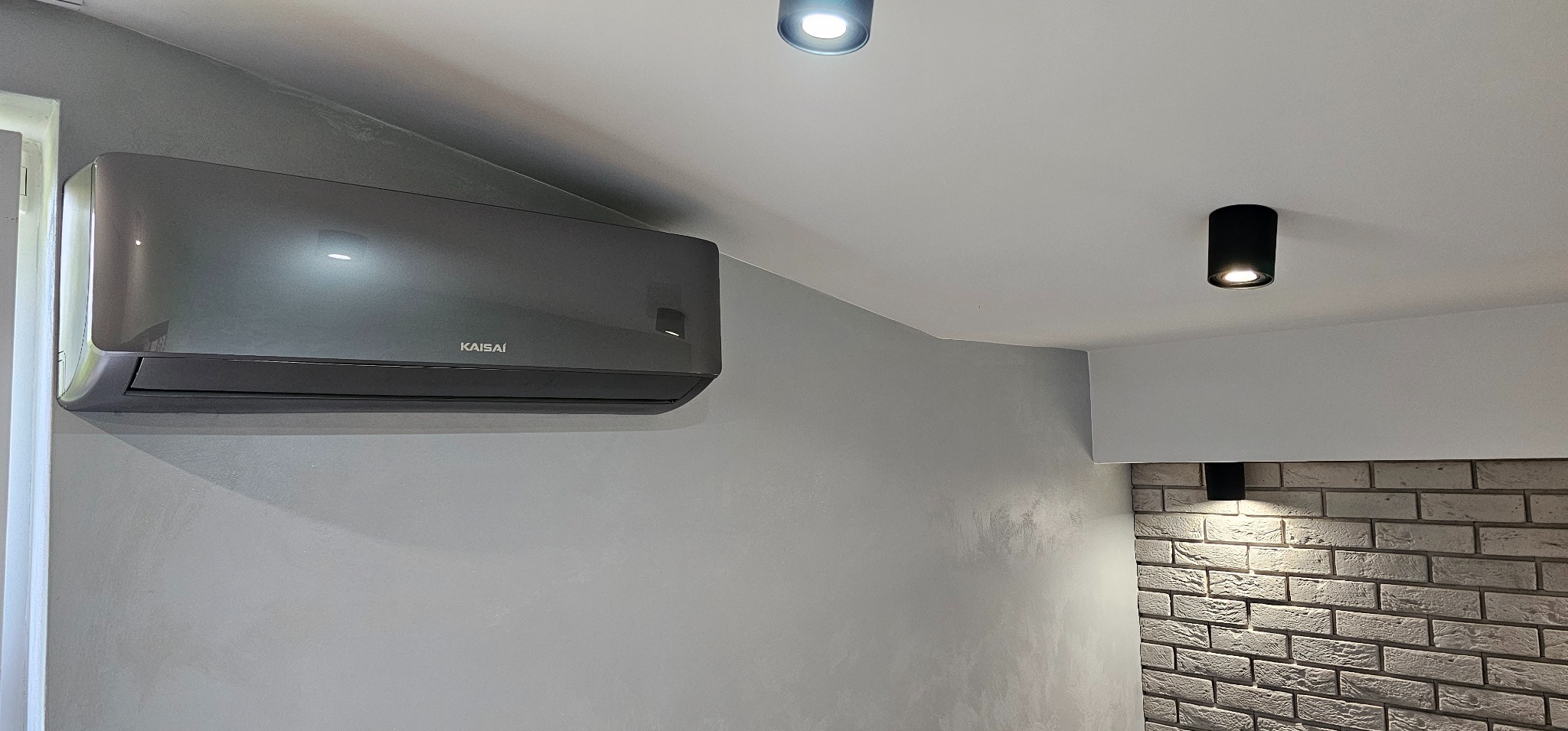 Szary klimatyzator Kaisai zamontowany na gładkiej, szarej ścianie, z widocznym fragmentem dekoracyjnej ściany z białej cegły i oświetleniem punktowym na suficie.