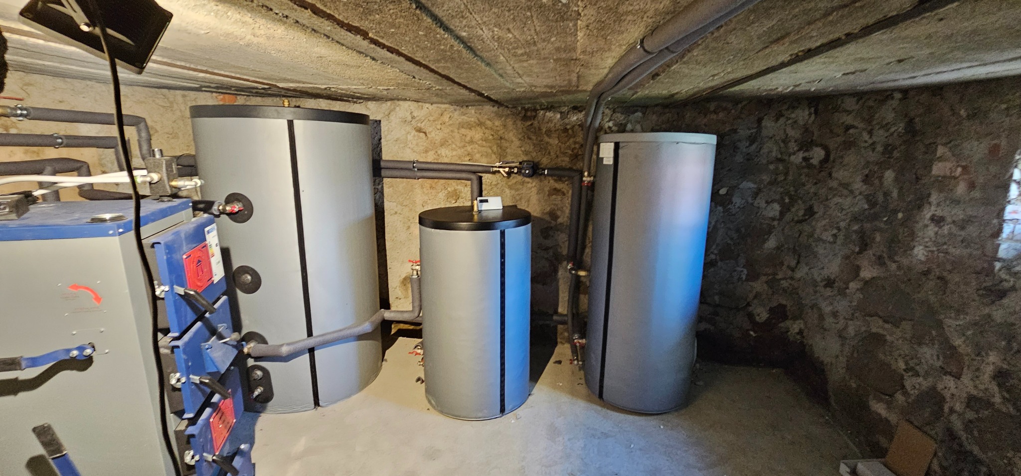 Instalacja buforów ciepła w kotłowni w Koźminku, z widocznymi rurami i elementami przyłączeniowymi, na tle surowej ściany z kamienia.