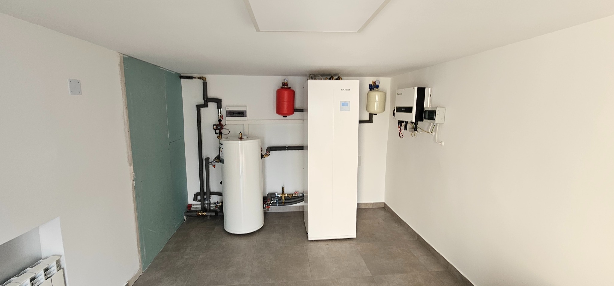 Wnętrze kotłowni z zainstalowaną białą pompą ciepła Kaisai, zasobnikiem wody, zbiornikiem wyrównawczym i inwerterem fotowoltaicznym Growatt na ścianie, widoczne rury instalacji grzewczej.