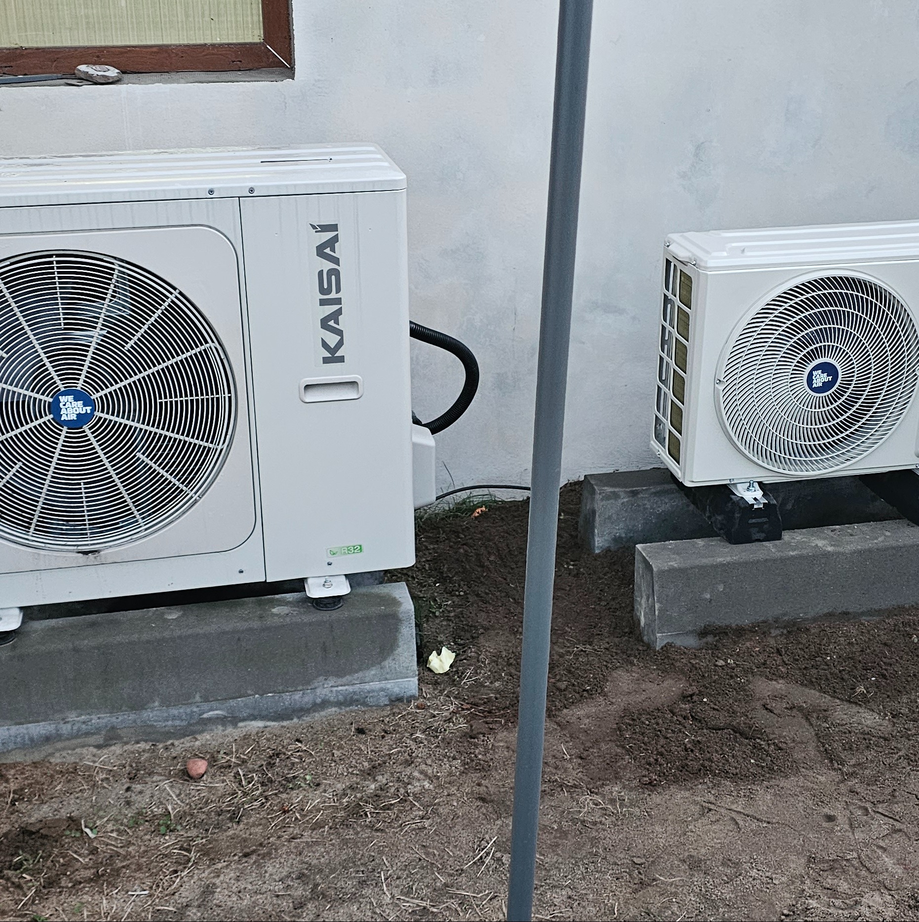 Dwie jednostki zewnętrzne klimatyzacji marki Kaisai, zamontowane na betonowych podstawach przy szarej ścianie budynku, z widocznymi elementami instalacji i nieporządkiem na ziemi.