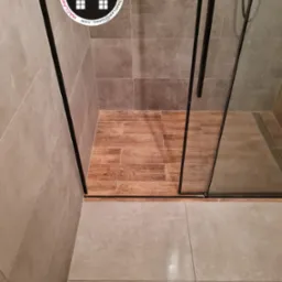 Wykończona kabina prysznicowa z brązowymi płytkami imitującymi drewno na podłodze, szklane drzwi z czarnymi ramami, oraz logo firmy glazurniczej w lewym górnym rogu.