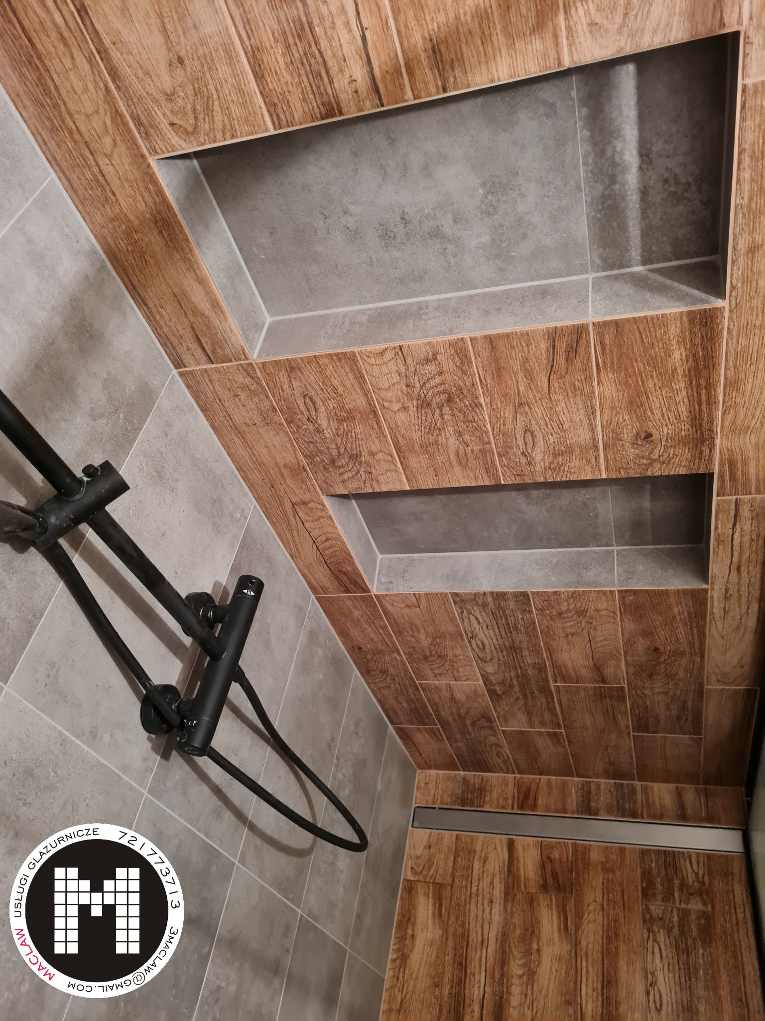 Wykończona łazienka z prysznicem typu walk-in, ściany i podłoga wyłożone płytkami imitującymi drewno, w ścianie wnęki wykończone szarą płytką, widoczny czarny zestaw prysznicowy.