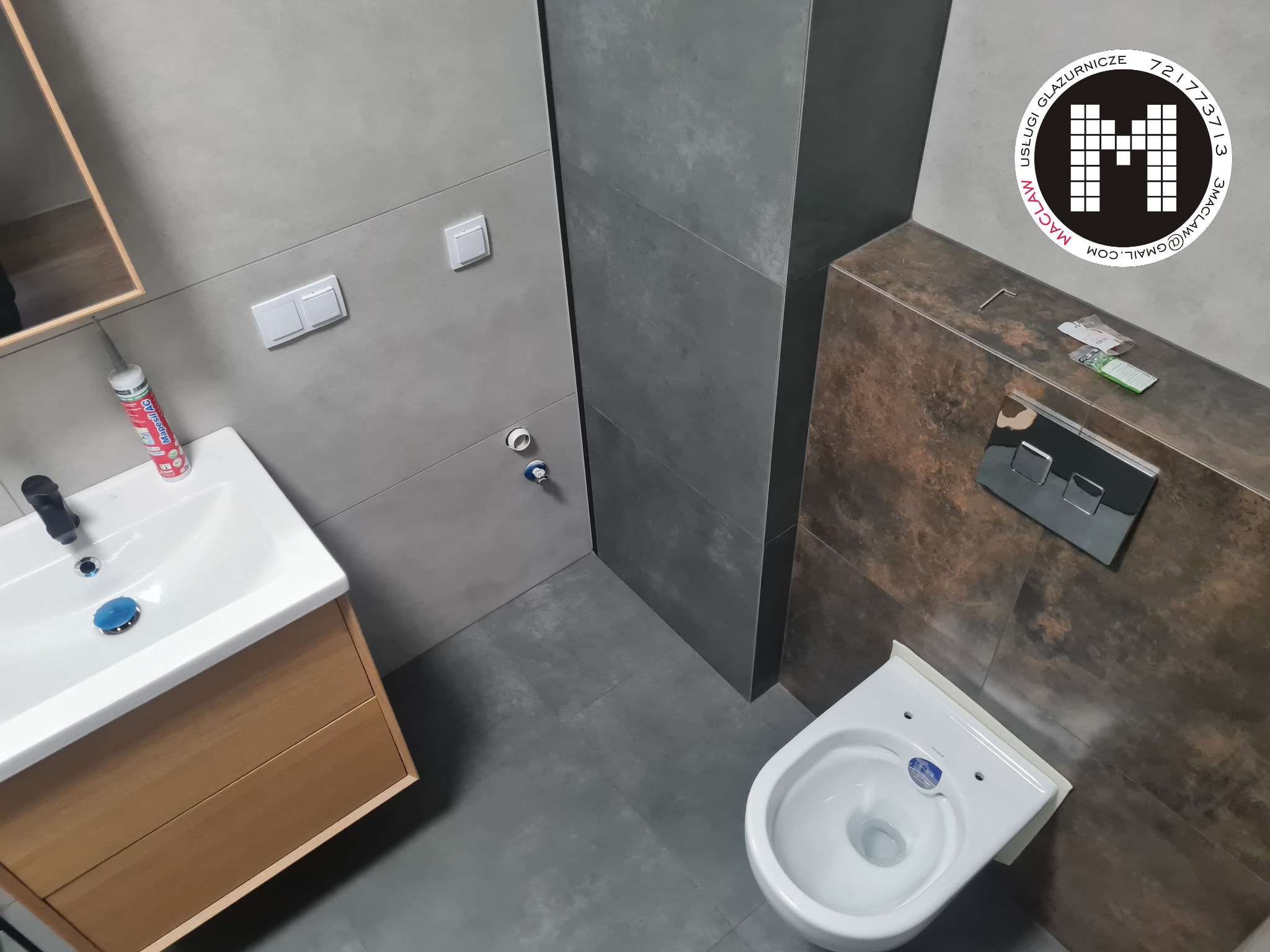 Nowoczesna łazienka z szarymi płytkami, drewnianą szafką pod umywalką i podwieszanym WC. Na brązowej ścianie widoczny panel sterujący spłuczką i logo firmy. Na blacie leżą narzędzia i dokumentacja.