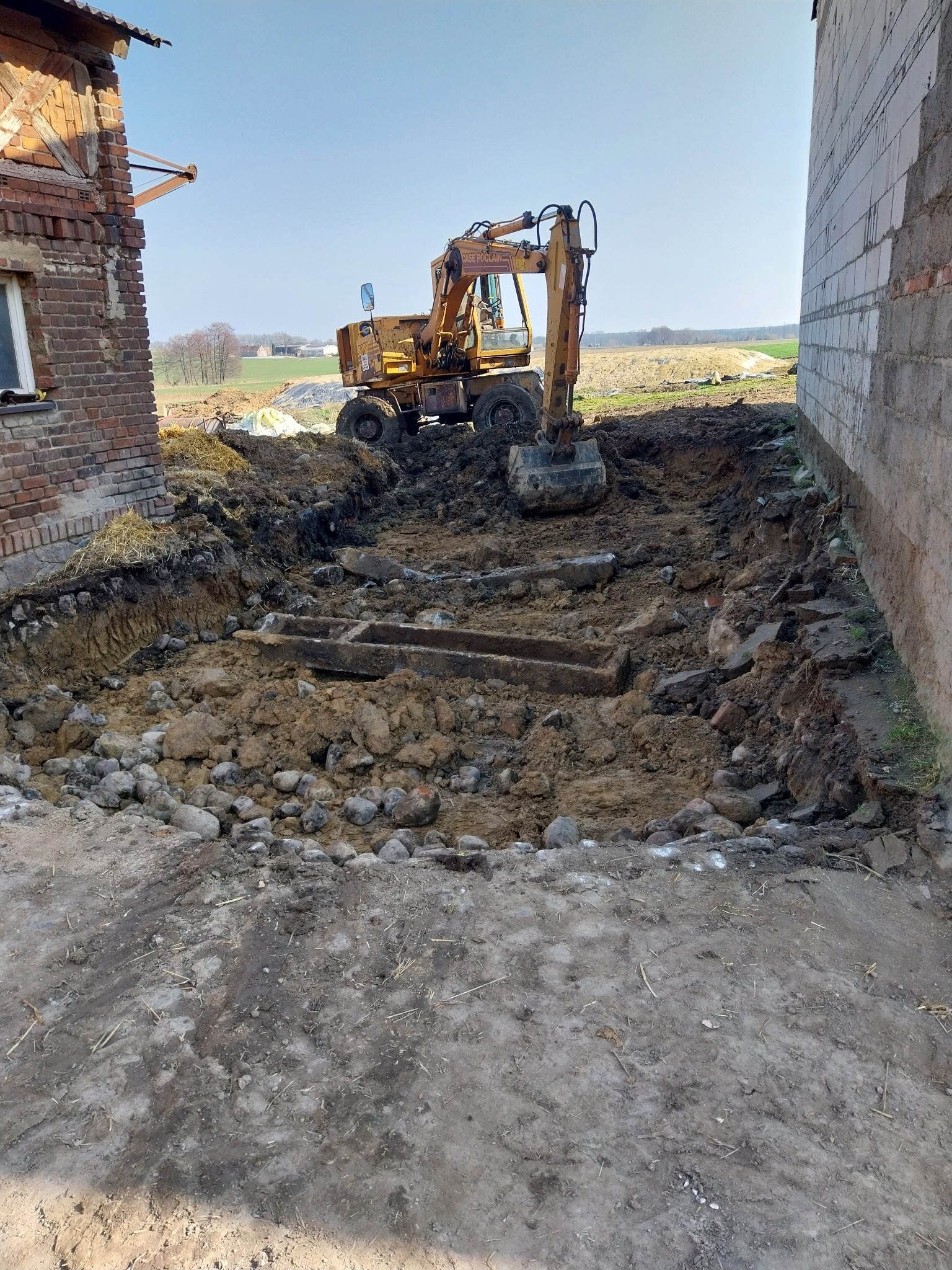 Żółta koparka wykonuje wykop fundamentów obok murowanego budynku we Wronkach, widoczne fragmenty starego fundamentu i kamienie.