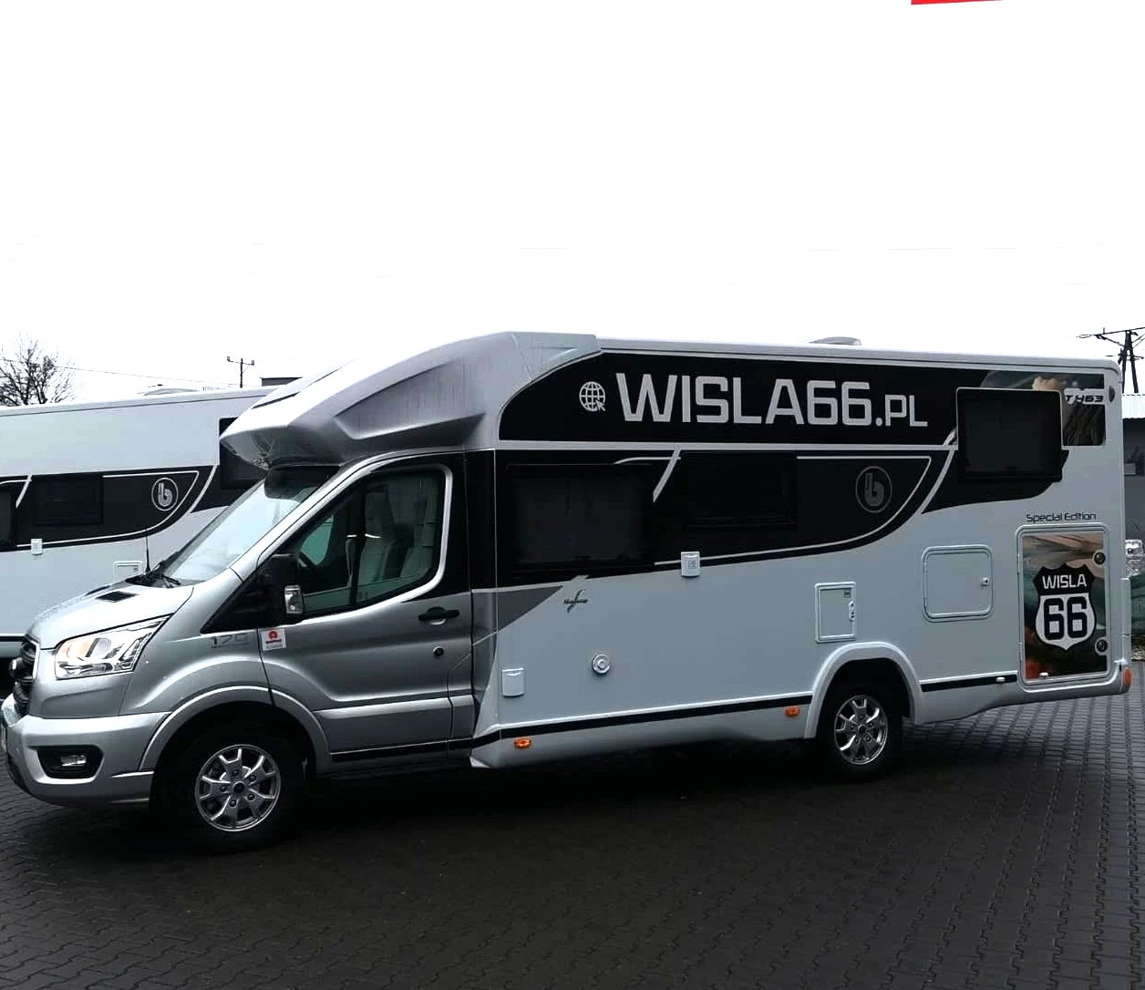 Srebrno-biały kamper Benimar Tessoro 463 z naklejką 'WISLA 66' na tylnych drzwiach, zaparkowany na mokrym bruku, widok z boku.