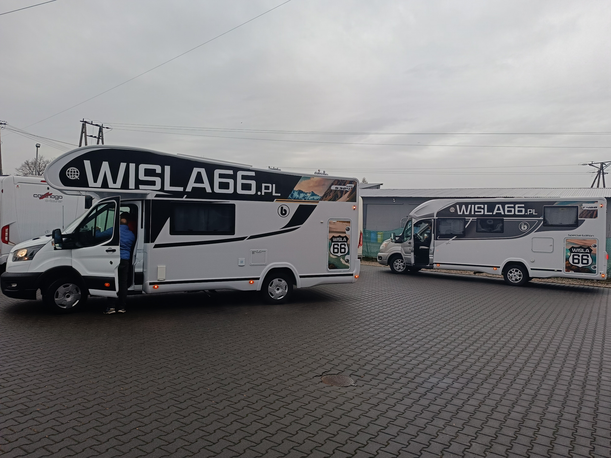 Dwa białe kampery z logo 'WISLA66.PL' na tle pochmurnego nieba i brukowanej powierzchni. Jeden z kamperów ma otwarte drzwi, z których wychodzi osoba w niebieskiej bluzie.