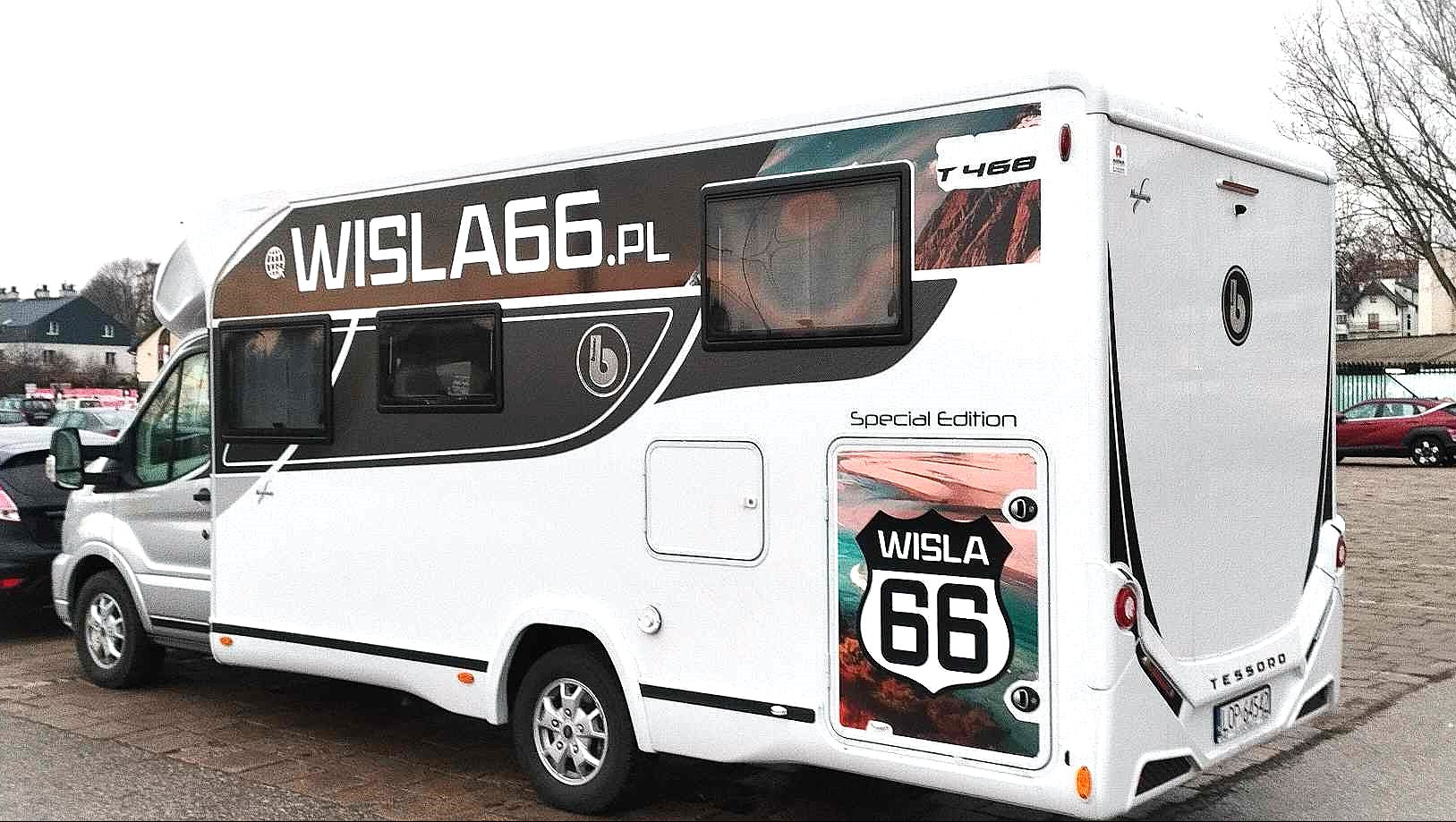 Biały kamper Benimar Tessoro 468 z grafiką WISLA66.PL na boku, zaparkowany na parkingu miejskim w pochmurny dzień. Widoczne okna z ciemnymi ramami i logo firmy na karoserii.