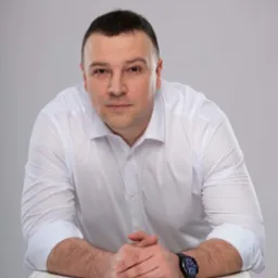 Portret mężczyzny w białej koszuli z zegarkiem, opierającego się o biały stół na jasnoszarym tle.