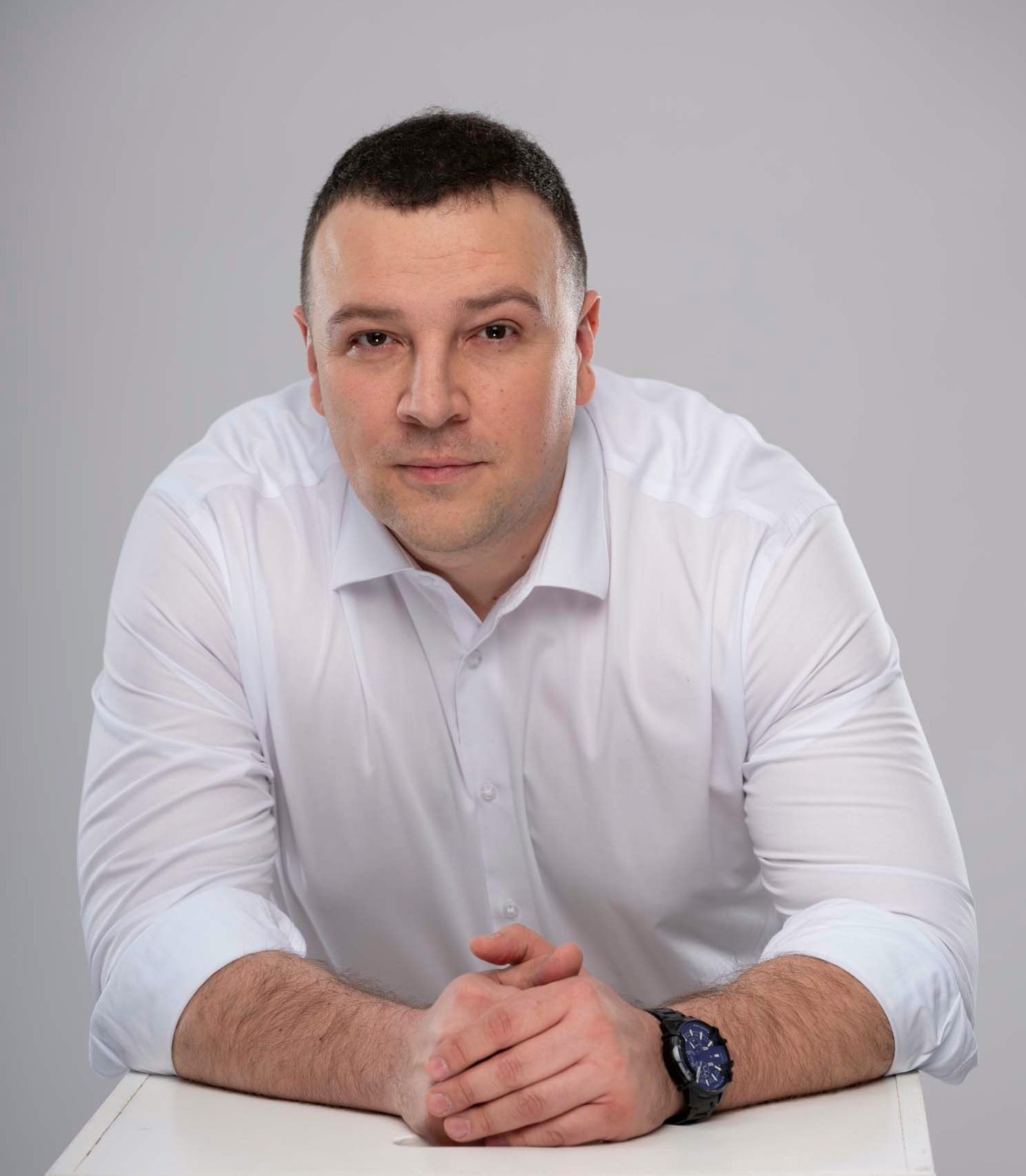 Portret mężczyzny w białej koszuli z zegarkiem, opierającego się o biały stół na jasnoszarym tle.