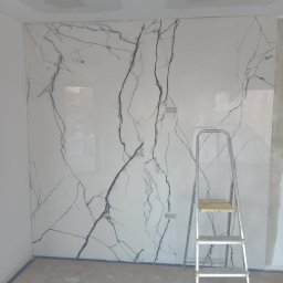 OPTIMUM GLOBAL PROSTA SPÓŁKA AKCYJNA - Ściana wykończona płytami imitującymi marmur Calacatta, z widoczną drabinką malarską, sugerująca prace wykończeniowe w trakcie realizacji.