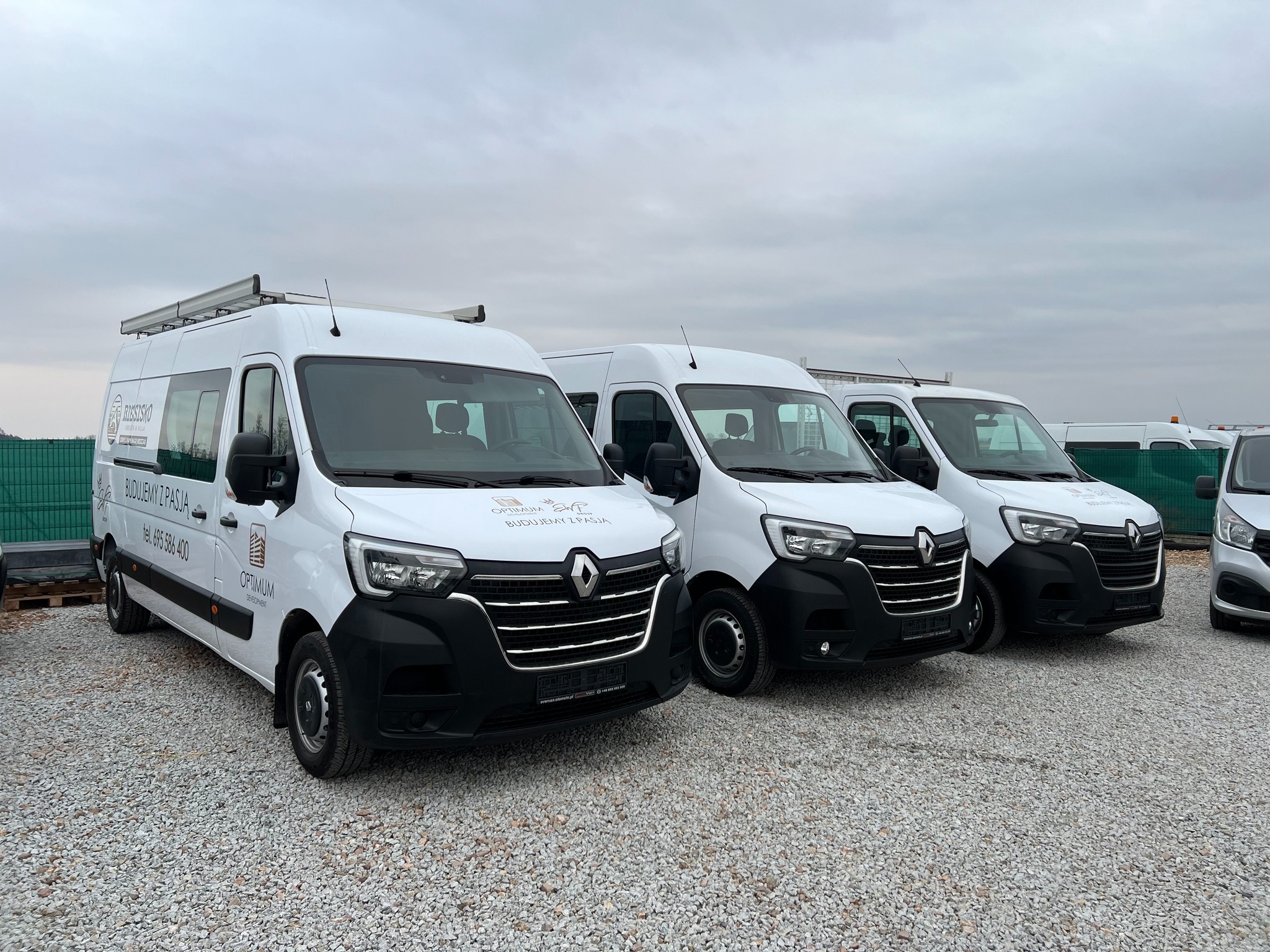 Flota białych samochodów dostawczych Renault Master z logo firmy budowlanej Rzeszisko Optimum Development, zaparkowanych na żwirowym placu pod pochmurnym niebem.
