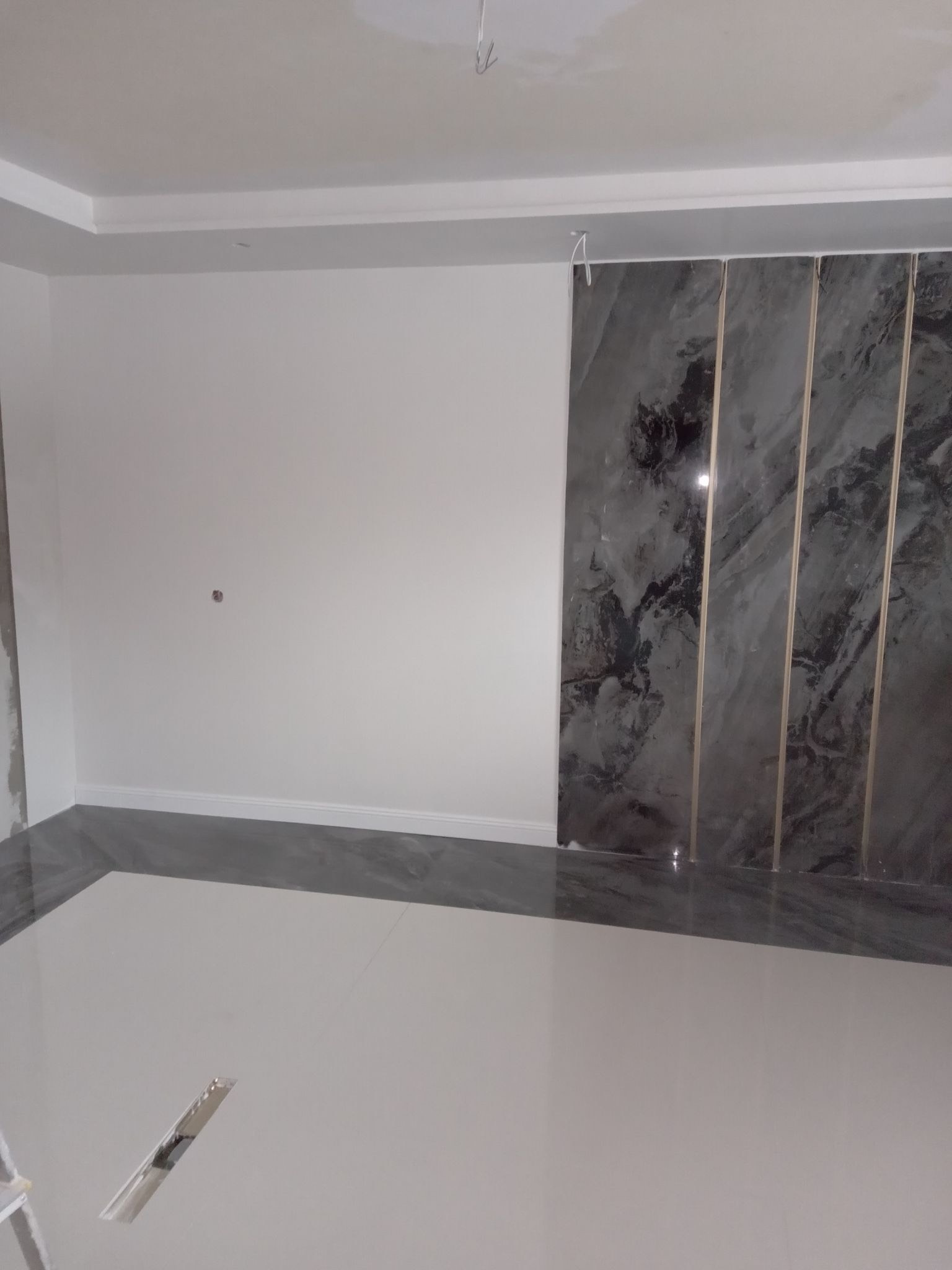 Wnętrze pokoju po remoncie, widoczna biała ściana, marmurowe panele z metalowymi listwami i błyszcząca podłoga w dwóch odcieniach szarości.