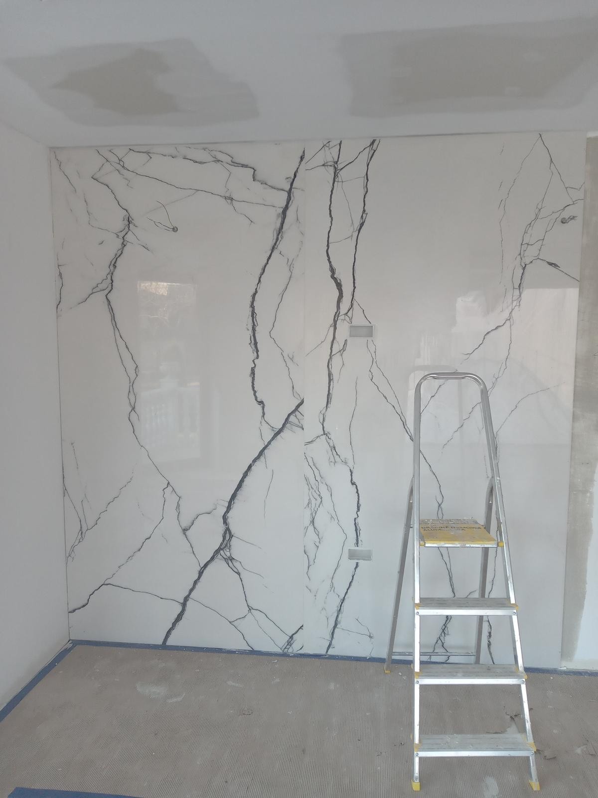 Ściana wykończona płytami imitującymi marmur Calacatta, z widoczną drabinką malarską, sugerująca prace wykończeniowe w trakcie realizacji.