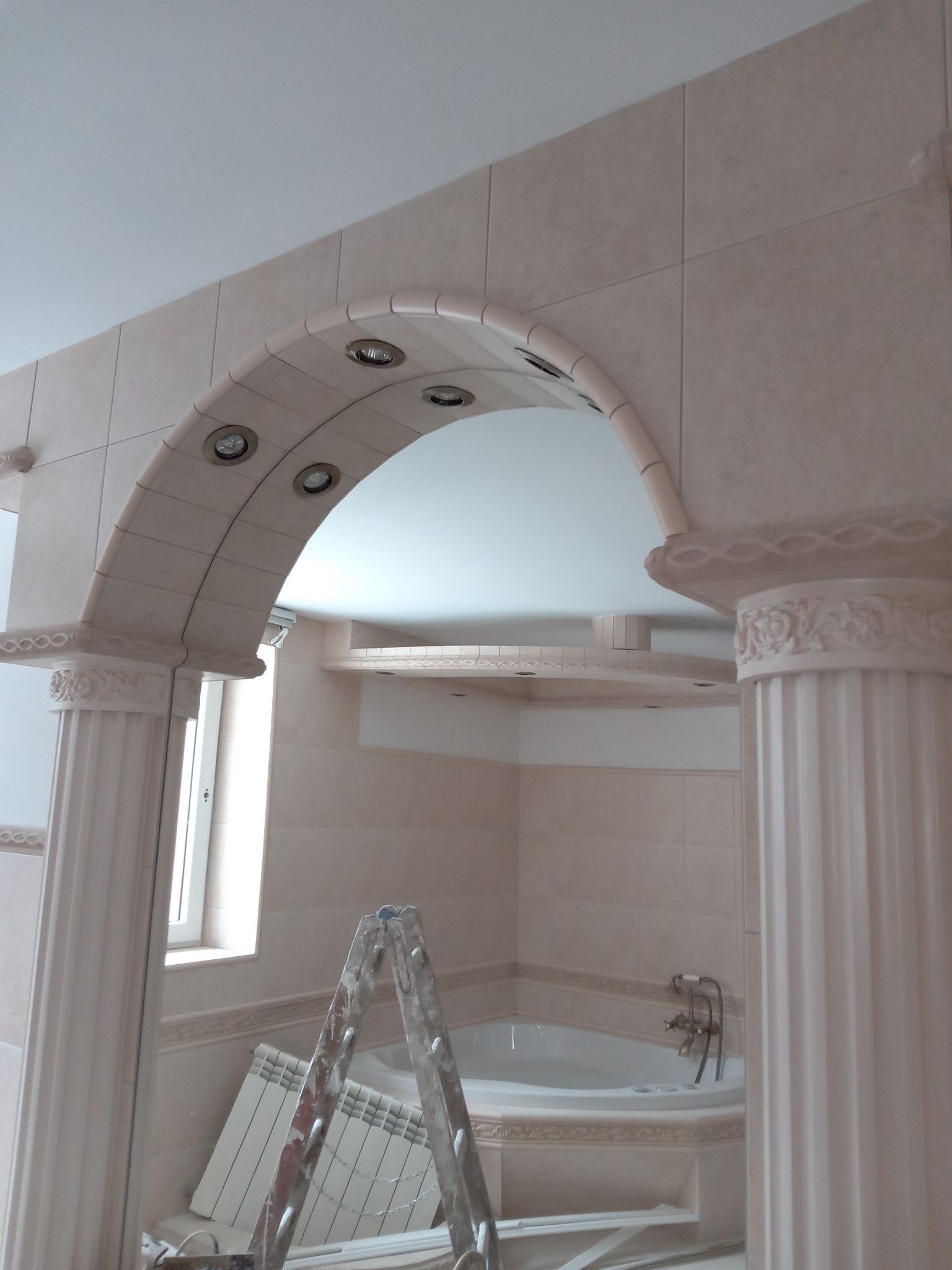 Wykończona łazienka z wanną, widoczny łuk z wbudowanym oświetleniem i kolumny z dekoracyjnymi elementami, częściowo zakryte drabiną malarską.