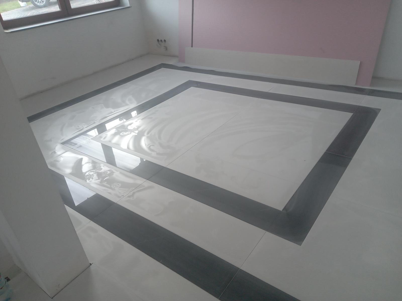 Układanie białych płytek podłogowych z ciemnoszarymi pasami dekoracyjnymi w Warszawskim wnętrzu, widoczne ślady świeżego kleju i okno w tle.