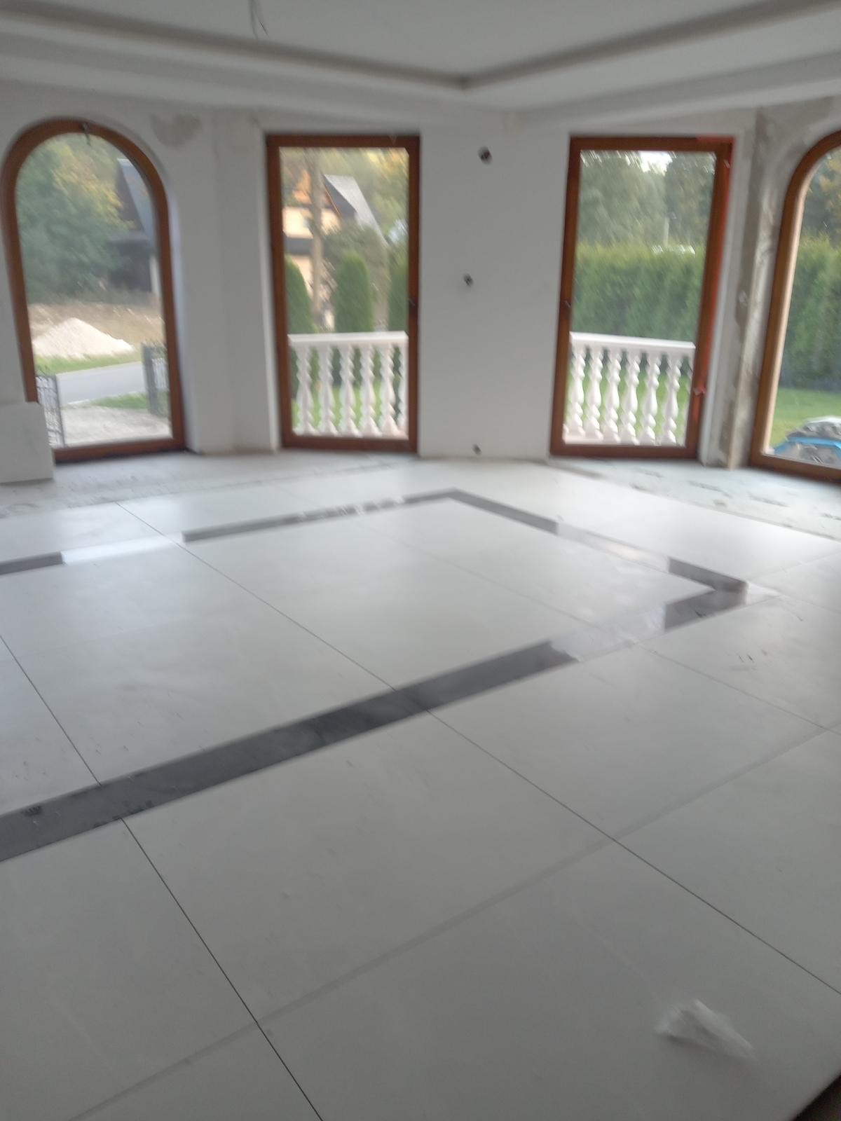 Wnętrze w trakcie prac wykończeniowych: białe płytki podłogowe z szarymi wstawkami układane w geometryczny wzór, okna z brązowymi ramami i widokiem na zieleń na zewnątrz, surowy stan ścian przed...