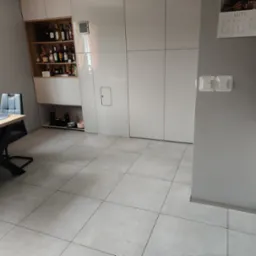 Jasne, kwadratowe płytki podłogowe w nowoczesnym wnętrzu z białymi szafkami, drewnianą półką z butelkami alkoholu i kalendarzem na ścianie.