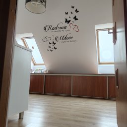 Paweł Gawron - Poddasze z oknami dachowymi, na ścianie dekoracyjny napis 'Rodzina tu zaczyna się życie a Miłość nigdy nie kończy' z motywem motyli i serc, widok zza otwartych drzwi.