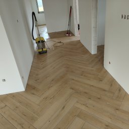Paweł Gawron - Ułożona podłoga z paneli w jodełkę w jasnym wnętrzu. Widoczny odkurzacz do wody i fragmenty ścian z instalacją elektryczną. Jasne, naturalne światło.