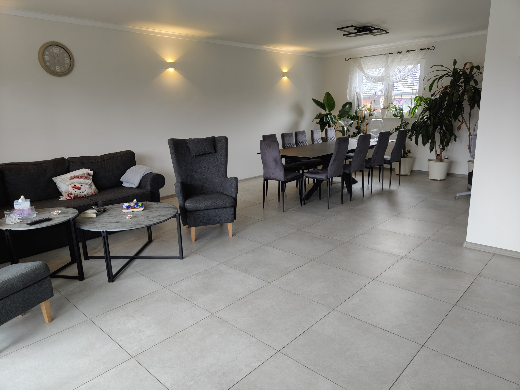 Jasne wnętrze salonu z szarymi płytkami podłogowymi, szarą sofą, fotelami i stołem jadalnym, oświetlone kinkietami i naturalnym światłem z okna, z roślinami doniczkowymi w tle.