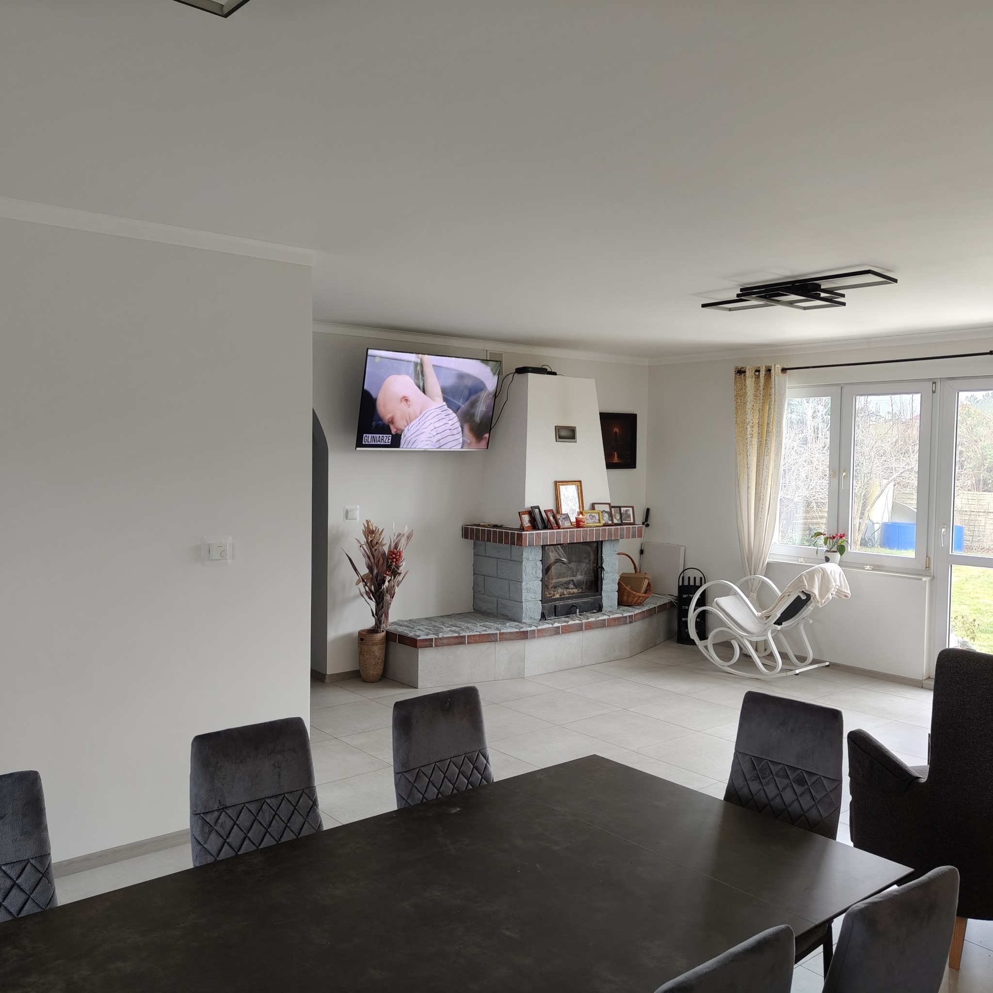 Przestronny salon z kominkiem obłożonym szarą cegłą, telewizorem nad kominkiem i dużym stołem z aksamitnymi krzesłami na pierwszym planie.
