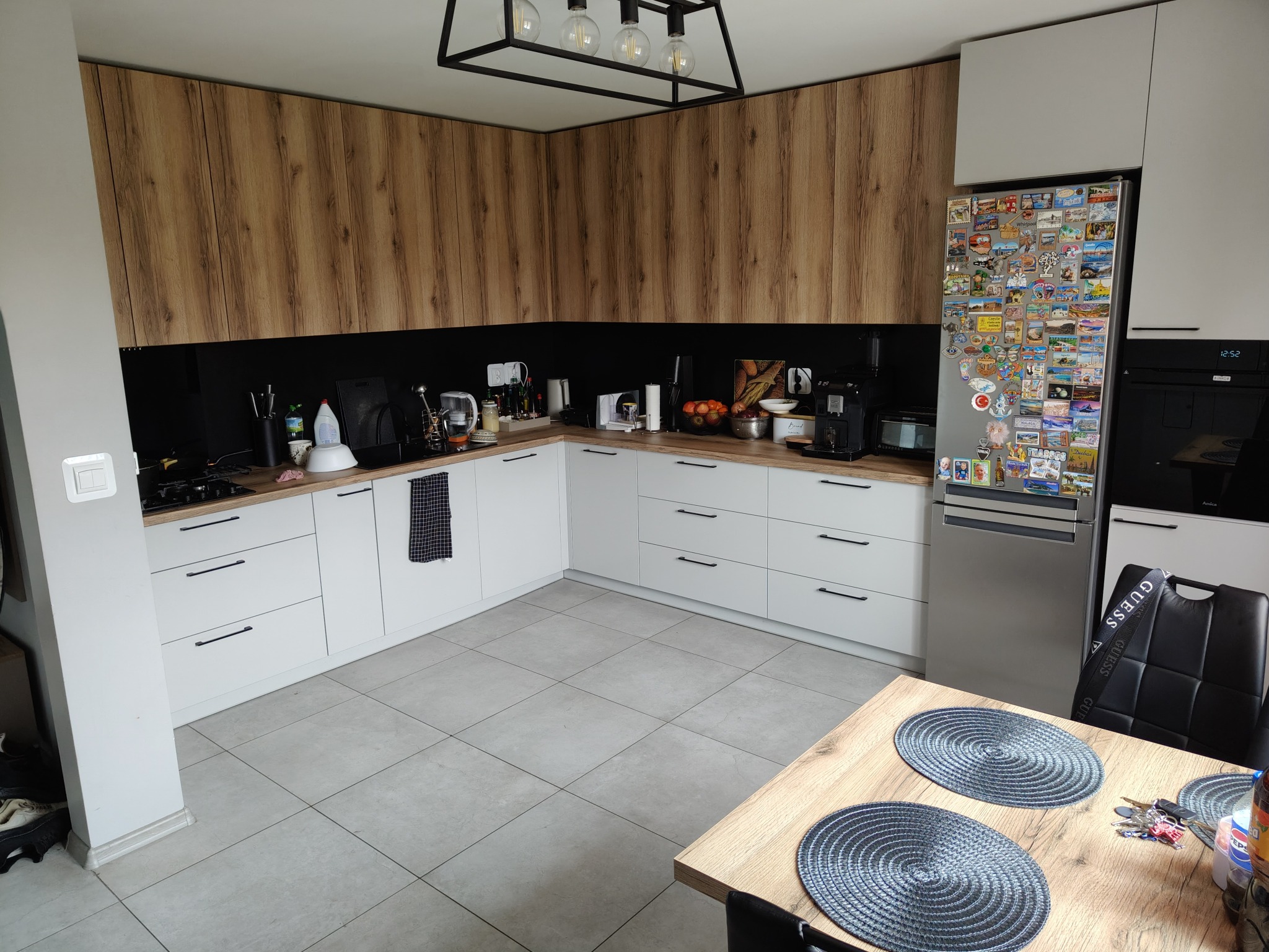 Nowoczesna kuchnia z białymi szafkami dolnymi i drewnopodobnymi górnymi, czarnym panelem nad blatem, szarą płytką podłogową i stołem z okrągłymi podkładkami; na lodówce widoczne magnesy z podróży.