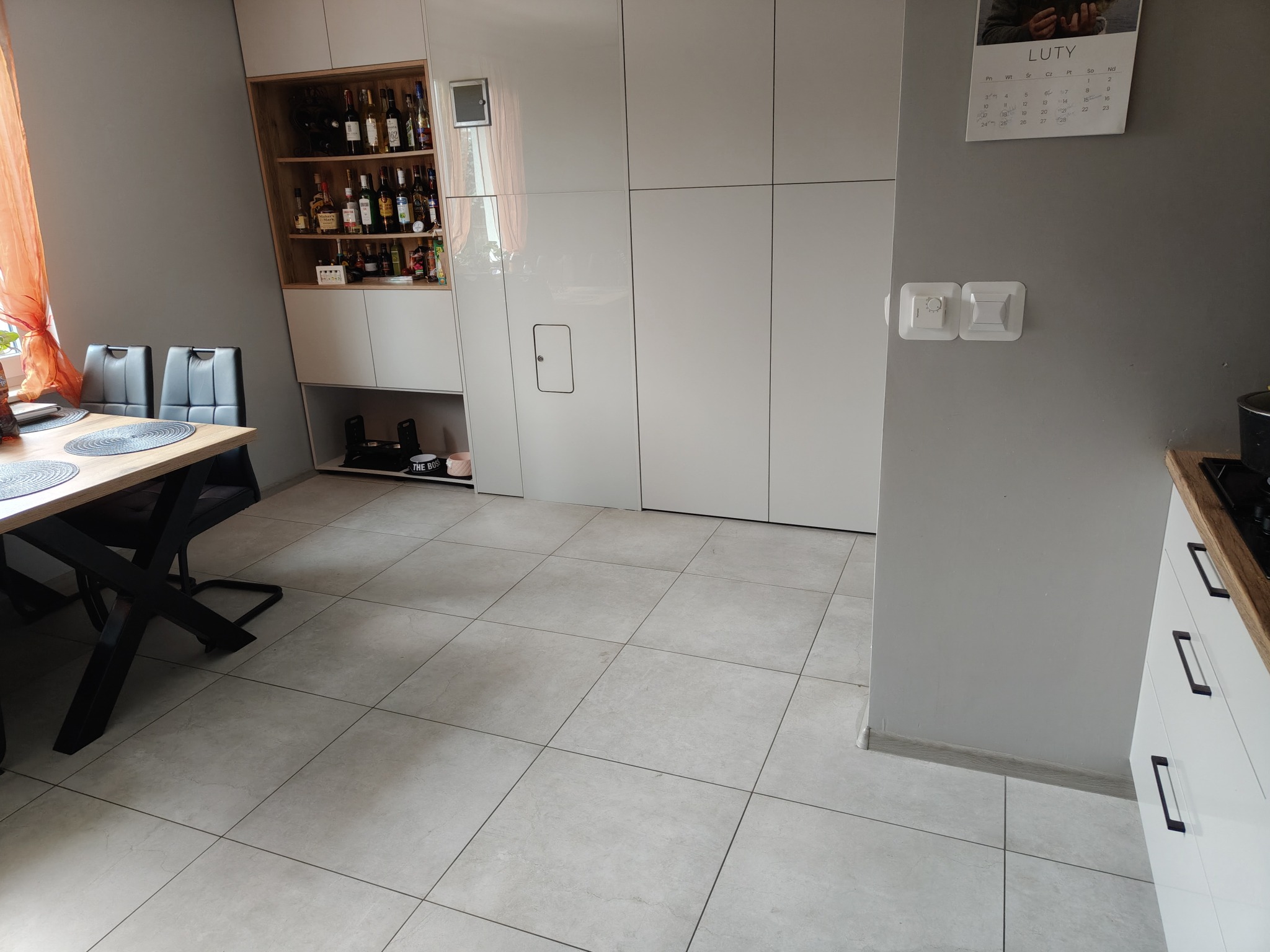 Jasne, kwadratowe płytki podłogowe w nowoczesnym wnętrzu z białymi szafkami, drewnianą półką z butelkami alkoholu i kalendarzem na ścianie.