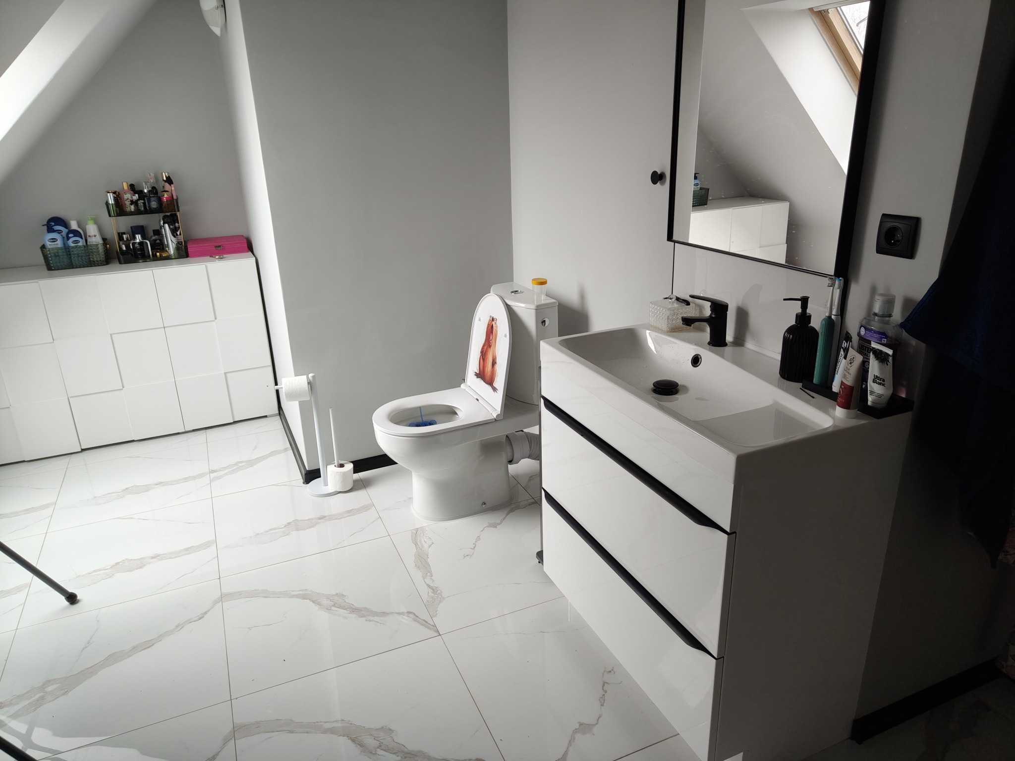 Nowoczesna łazienka na poddaszu z białą armaturą, marmurową podłogą i toaletą z humorystyczną naklejką na desce sedesowej.