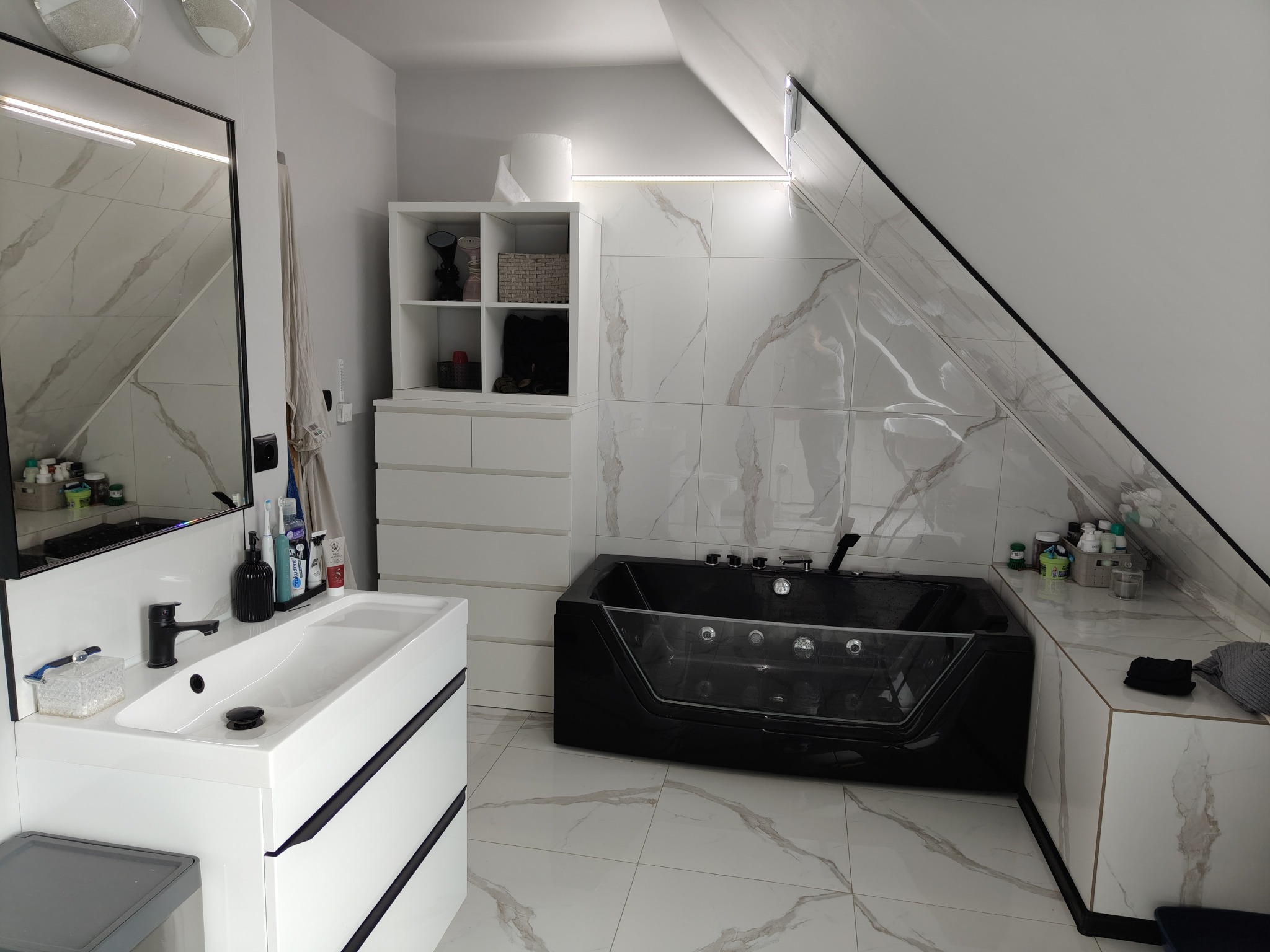 Nowoczesna łazienka na poddaszu z czarną wanną z hydromasażem, białą umywalką z czarną armaturą i marmurowymi płytkami na ścianach i podłodze.