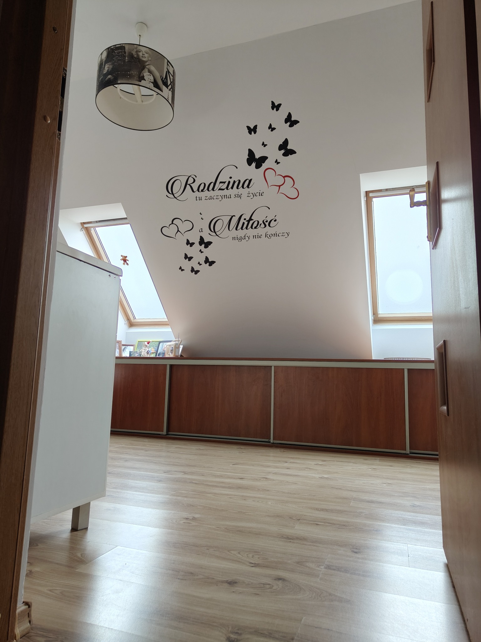 Poddasze z oknami dachowymi, na ścianie dekoracyjny napis 'Rodzina tu zaczyna się życie a Miłość nigdy nie kończy' z motywem motyli i serc, widok zza otwartych drzwi.