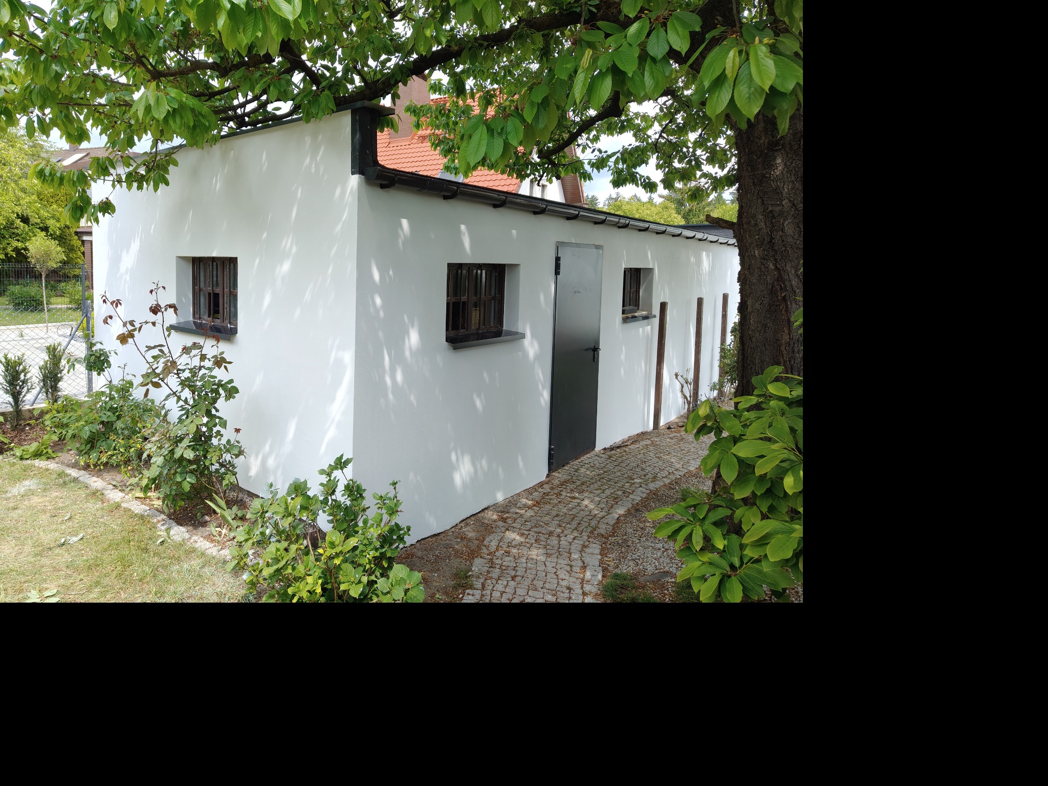 Biała, otynkowana ściana budynku z ciemnymi oknami i drzwiami, otoczona zielenią. Ścieżka z kostki brukowej prowadzi wzdłuż ściany. Widoczne cienie drzew.