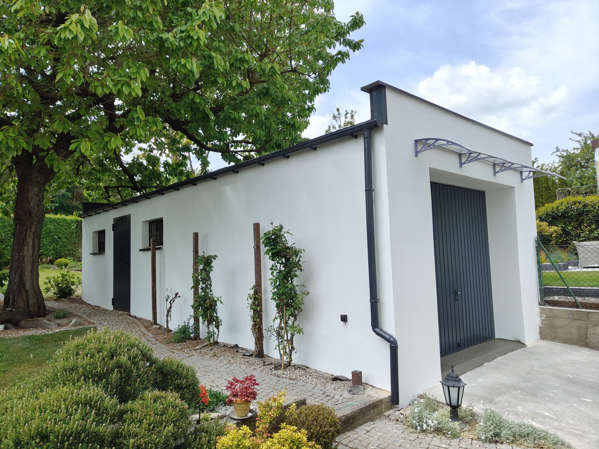 Biały budynek gospodarczy z szarą bramą garażową i zadaszeniem, otoczony zielenią i kostką brukową. Widoczne okna i rynna. Ujęcie w słoneczny dzień.