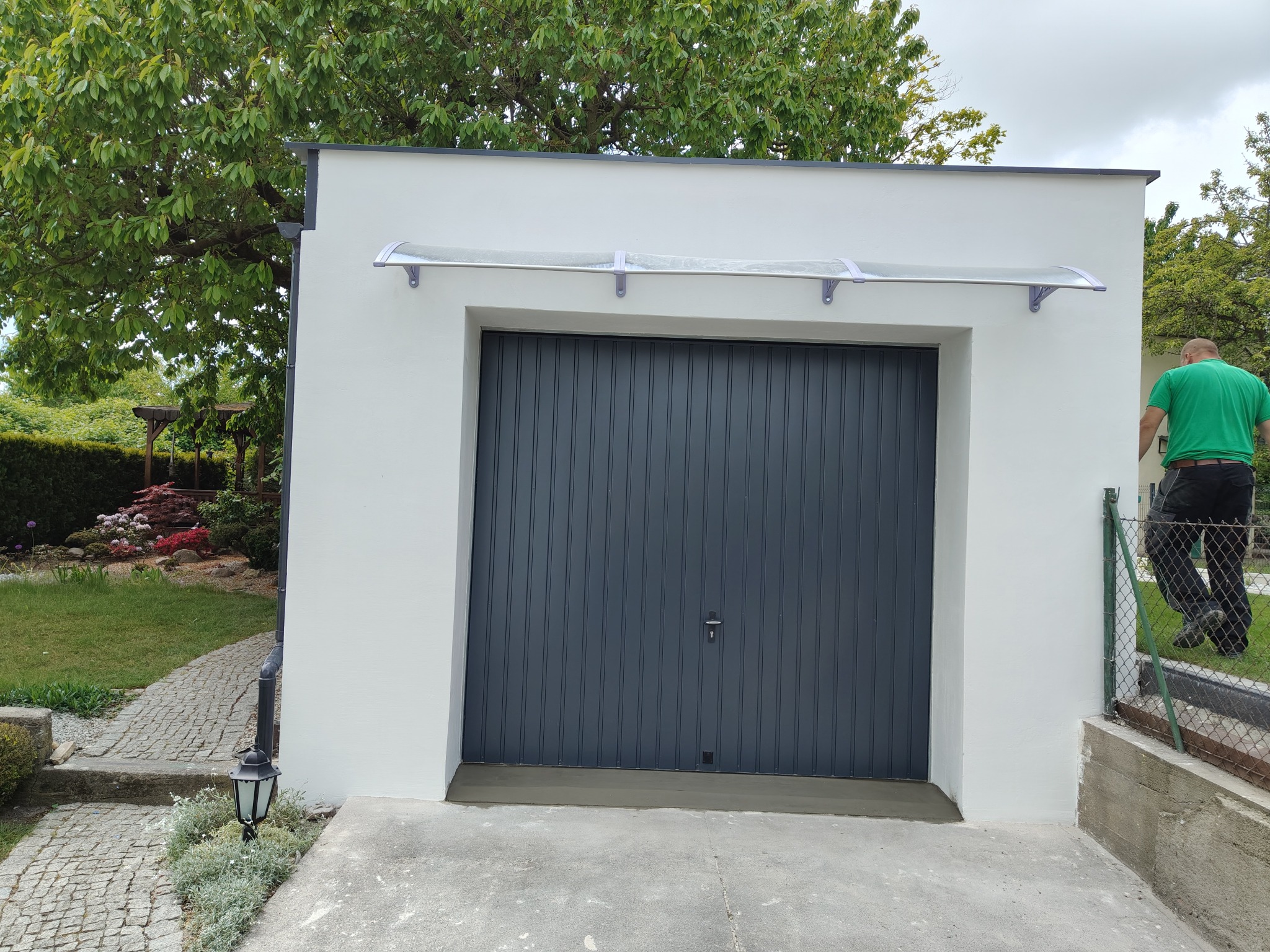 Biały garaż z szarą bramą segmentową i przezroczystym zadaszeniem. Świeżo pomalowana elewacja, widoczny fragment ogrodu i ścieżki z kostki brukowej. Osoba w tle.